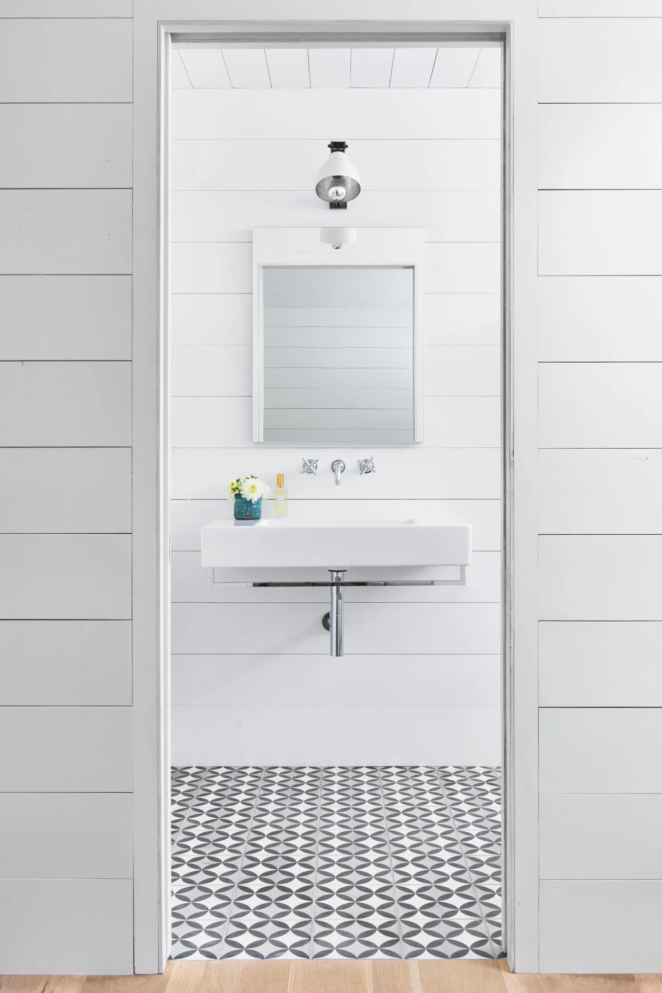 A peek inside a brightly lit bathroom with white and grey geometric tiling, a clean sink, and square mirror.