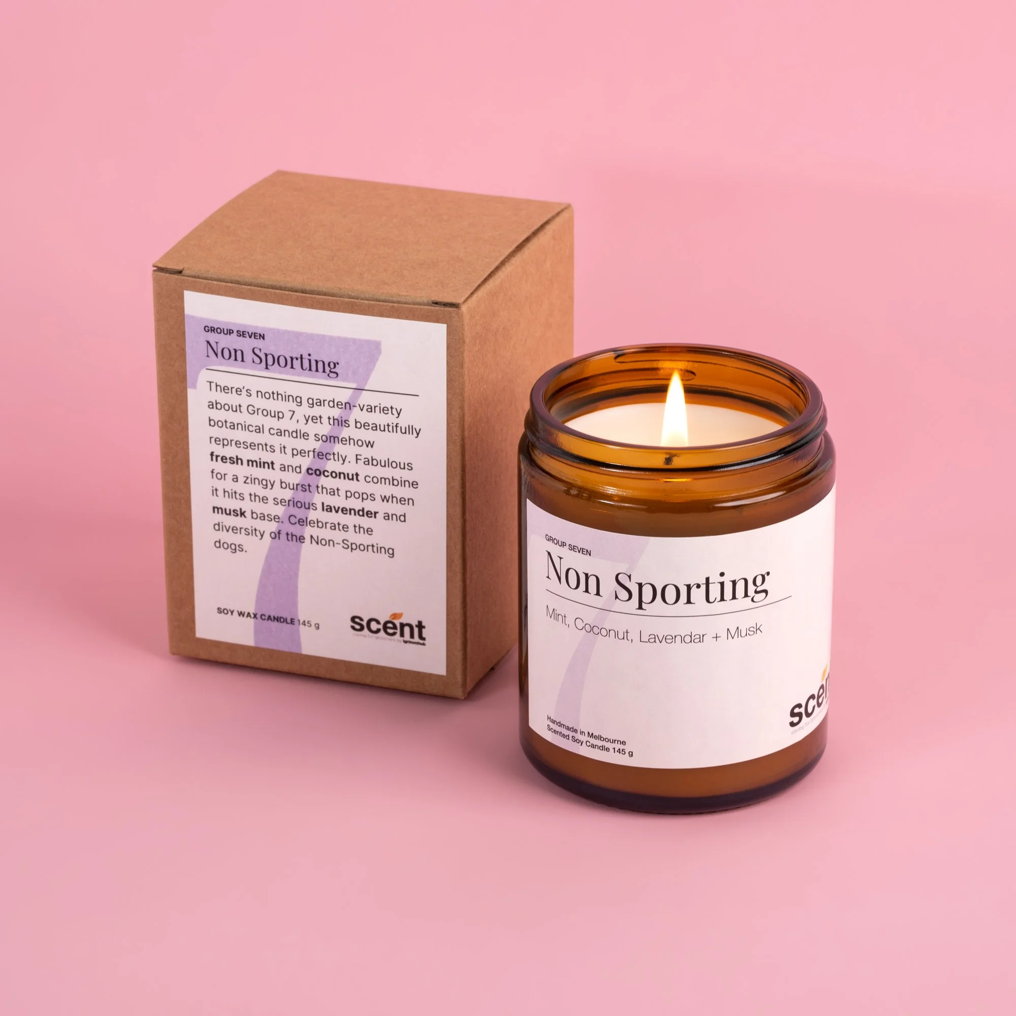 A lit brown glass candle with a white label reading 'Non Sporting' and a cardboard box with similar labeling, both placed on a pink surface.