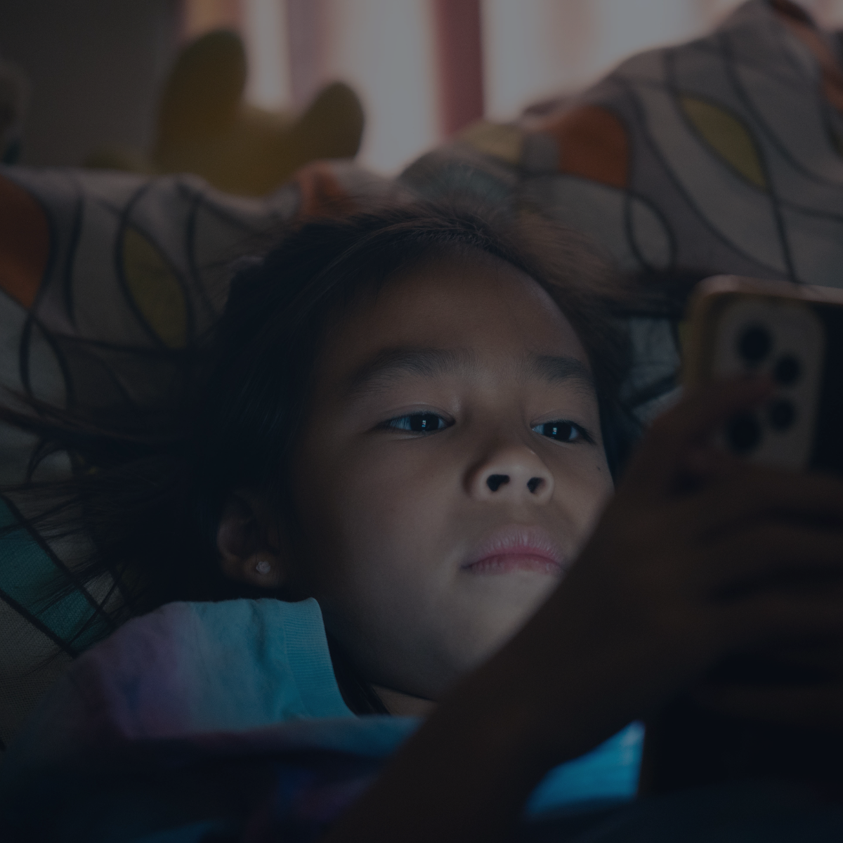 A young girl lying on her back in bed, looking at her phone. She has dark hair, a calm expression, and is wearing a light blue shirt. The bed has patterned sheets, and there is a potted plant in the background.
