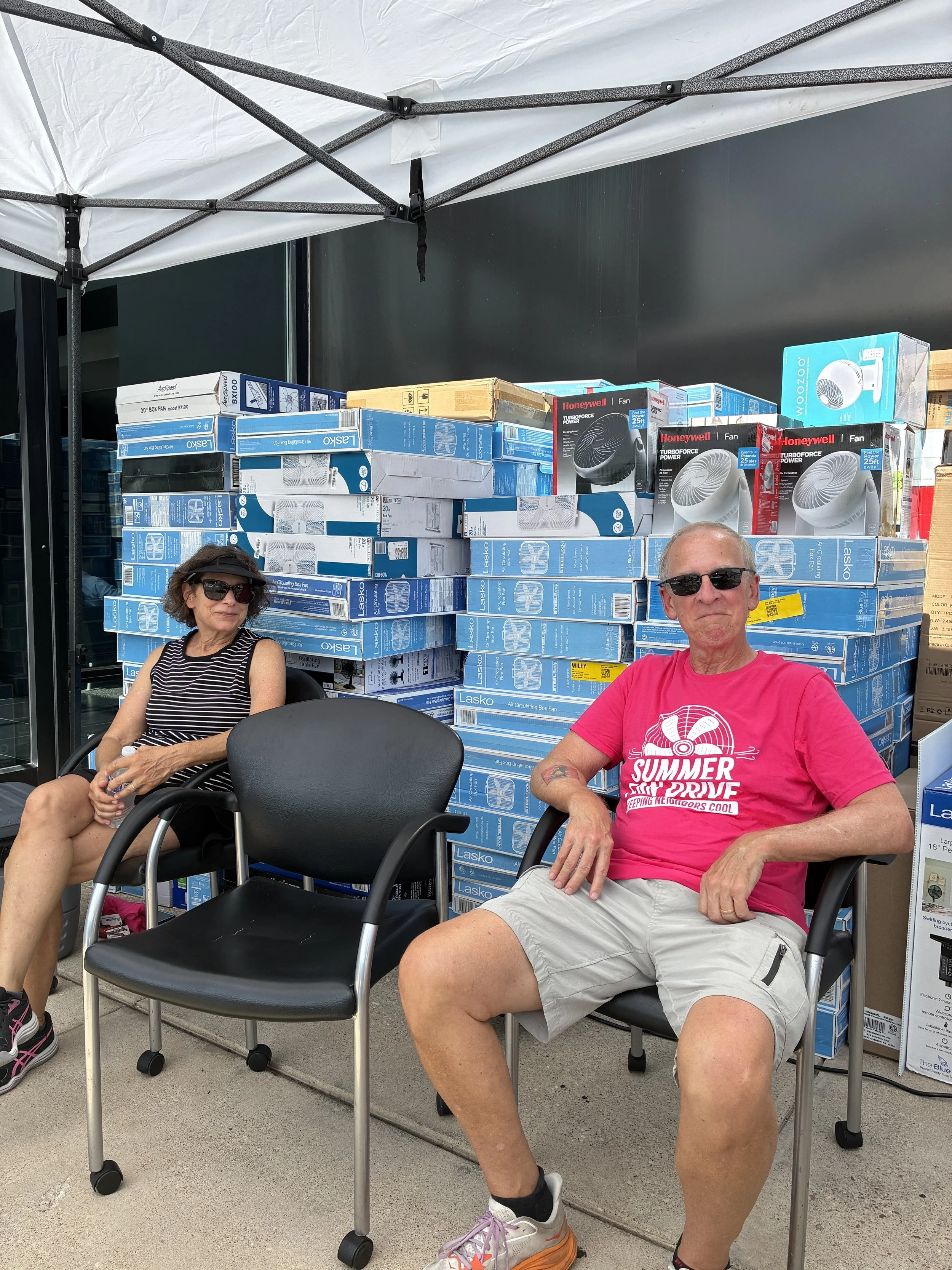 2 people sitting in chairs in front of stacks of box fans
