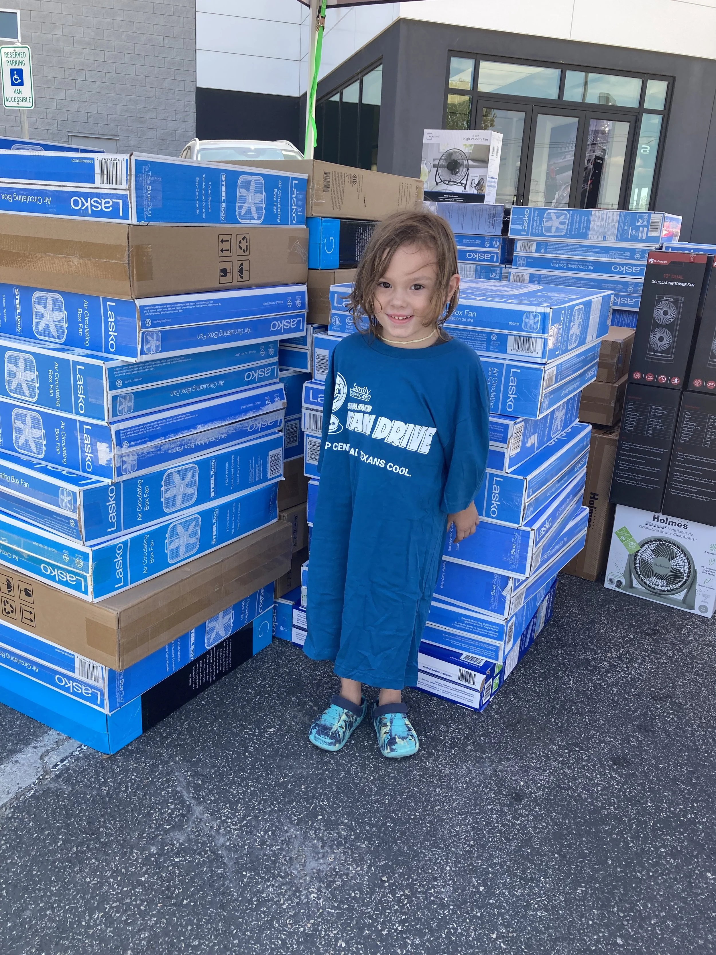 A kid wearing a Summer Fan Drive shirt standing by a stack of fans