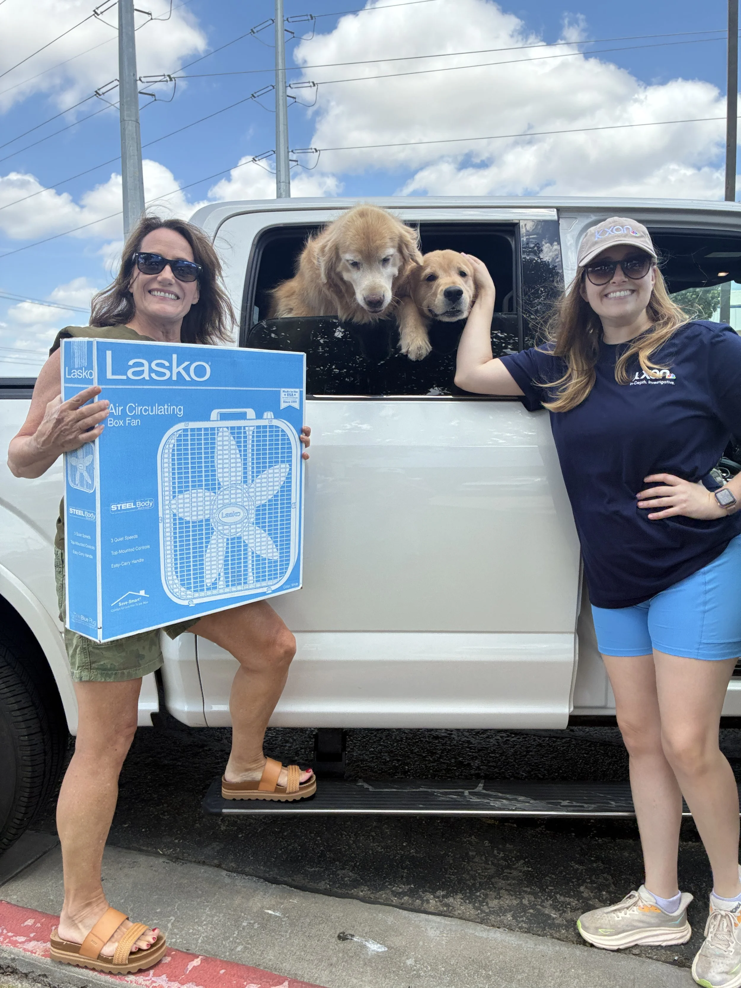 2 people standing by a car with two dogs in the window. One person is holding a box fan.