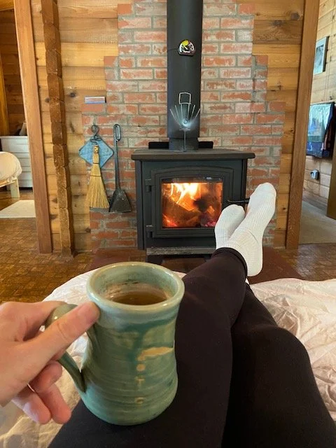Sarah holding a mug of tea with her legs crossed out in front of a wood burning stove
