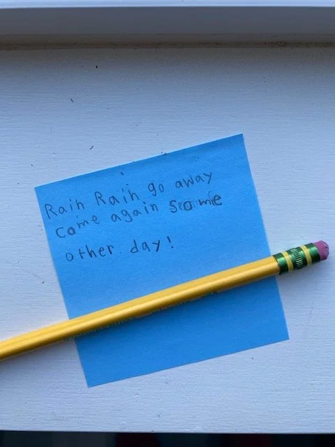 A blue Post-It note with children's writing that says "Rain, rain go away come again some other day!"