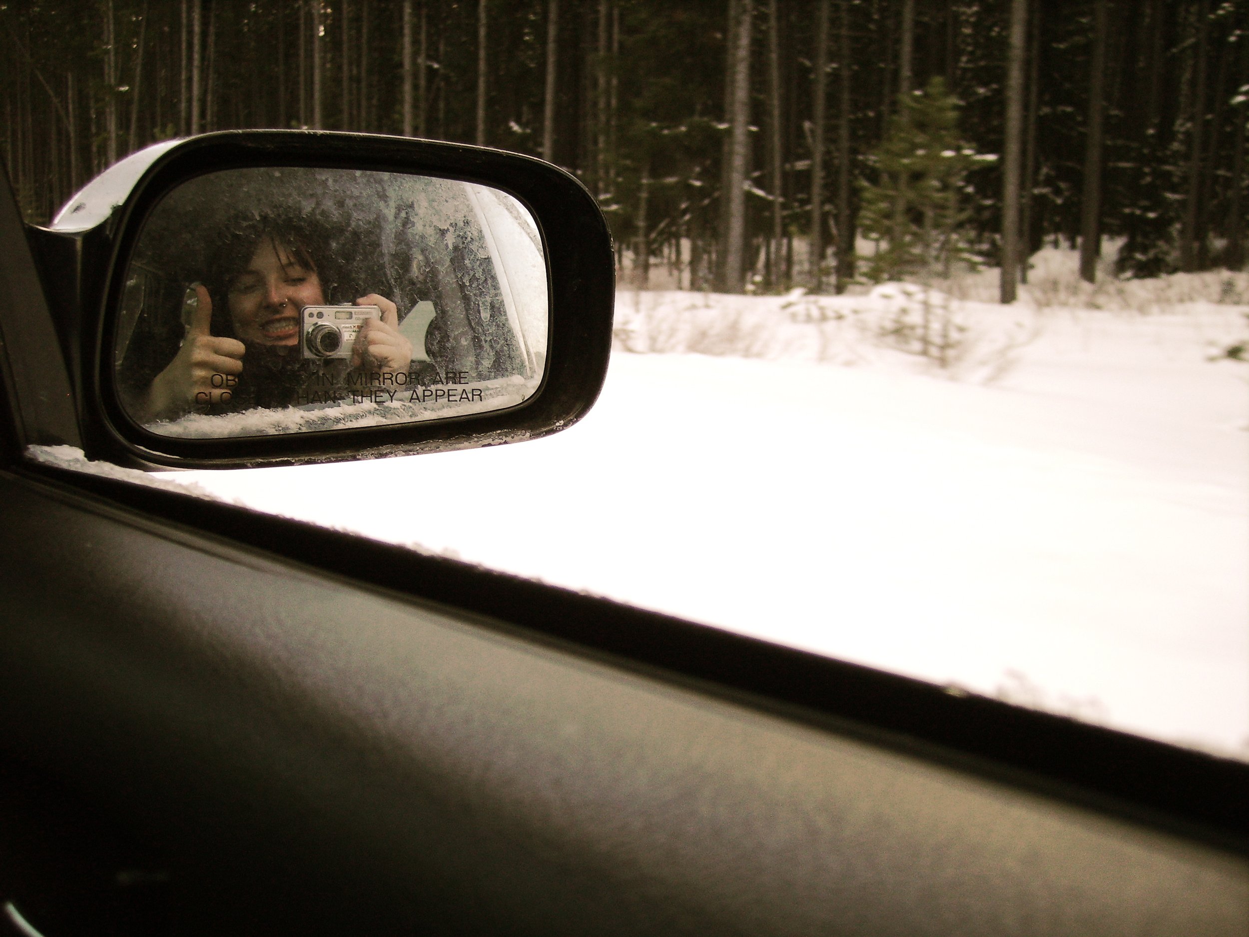 Sarah's reflection in a car side mirror holding a camera and giving a thumbs up with snow and trees out the window
