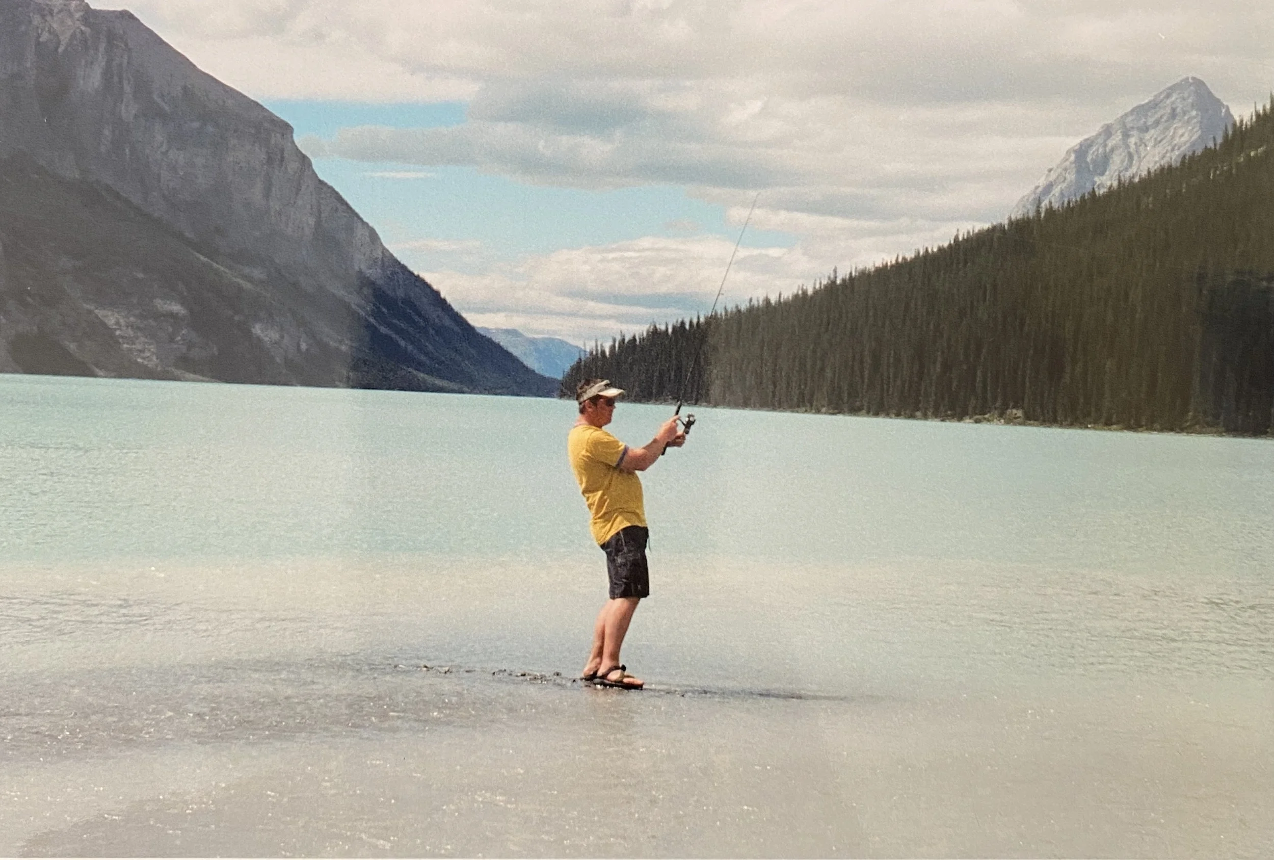 A man standing in shallow water with a fishing rod with beautiful mountains and a blue lake behind him