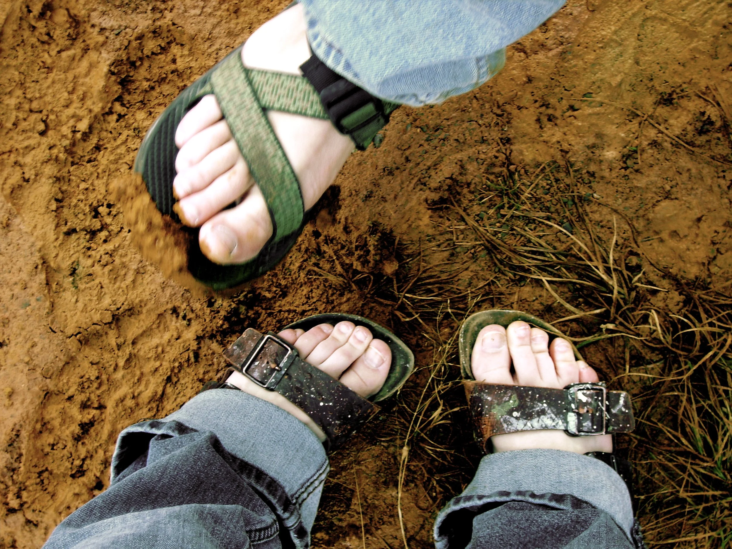 Sarah's feet in birkenstocks and a large man's foot in a Teva sandal on muddy earth