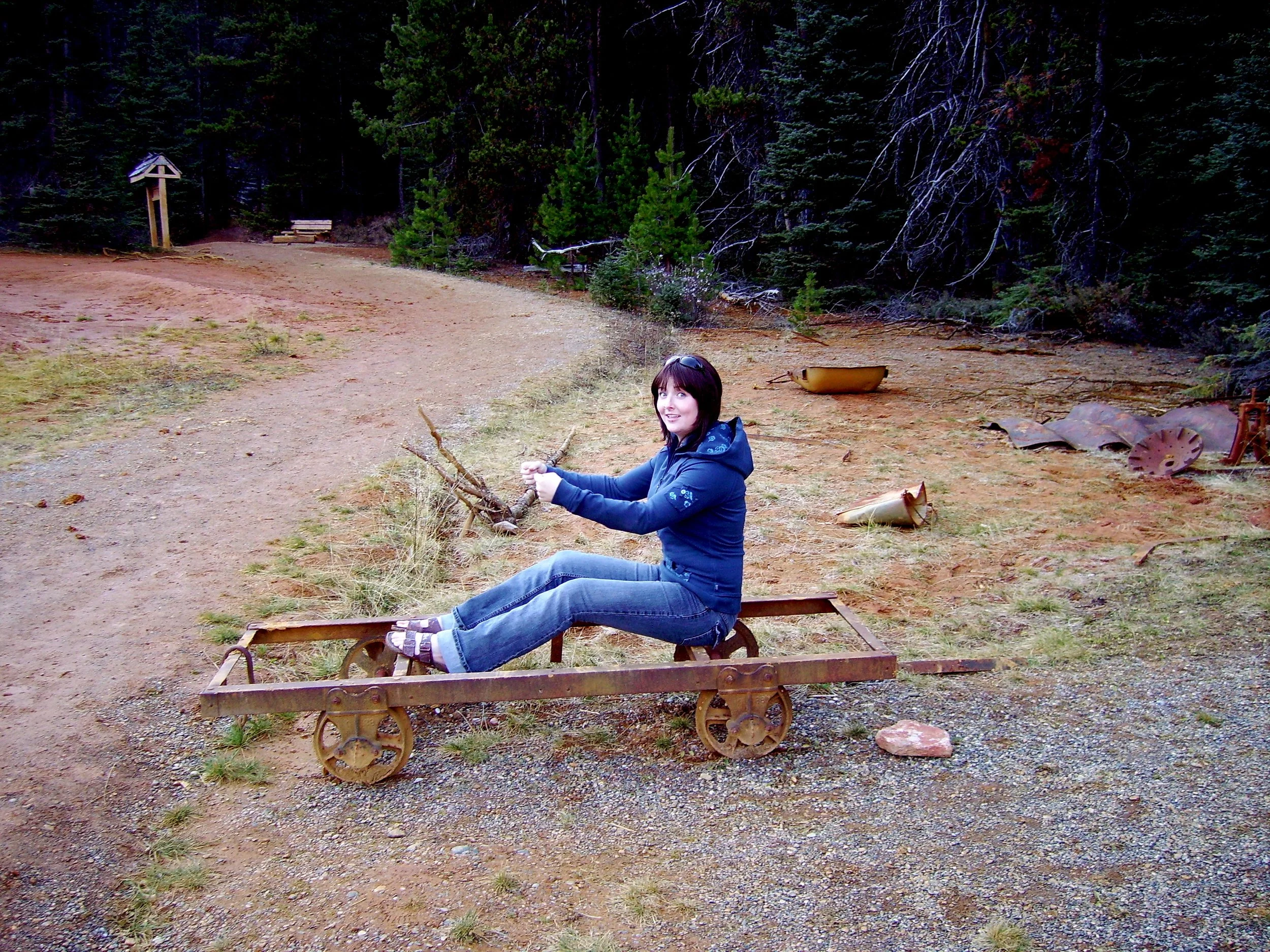 Sarah awkwardly sitting on a rusted mining cart pretending to drive it by a forest