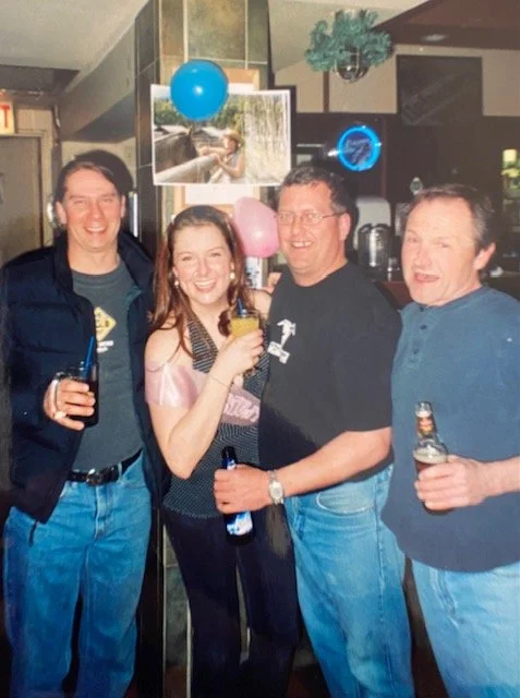 Sarah with her arms around 3 older men holding bottles of beer in a bar