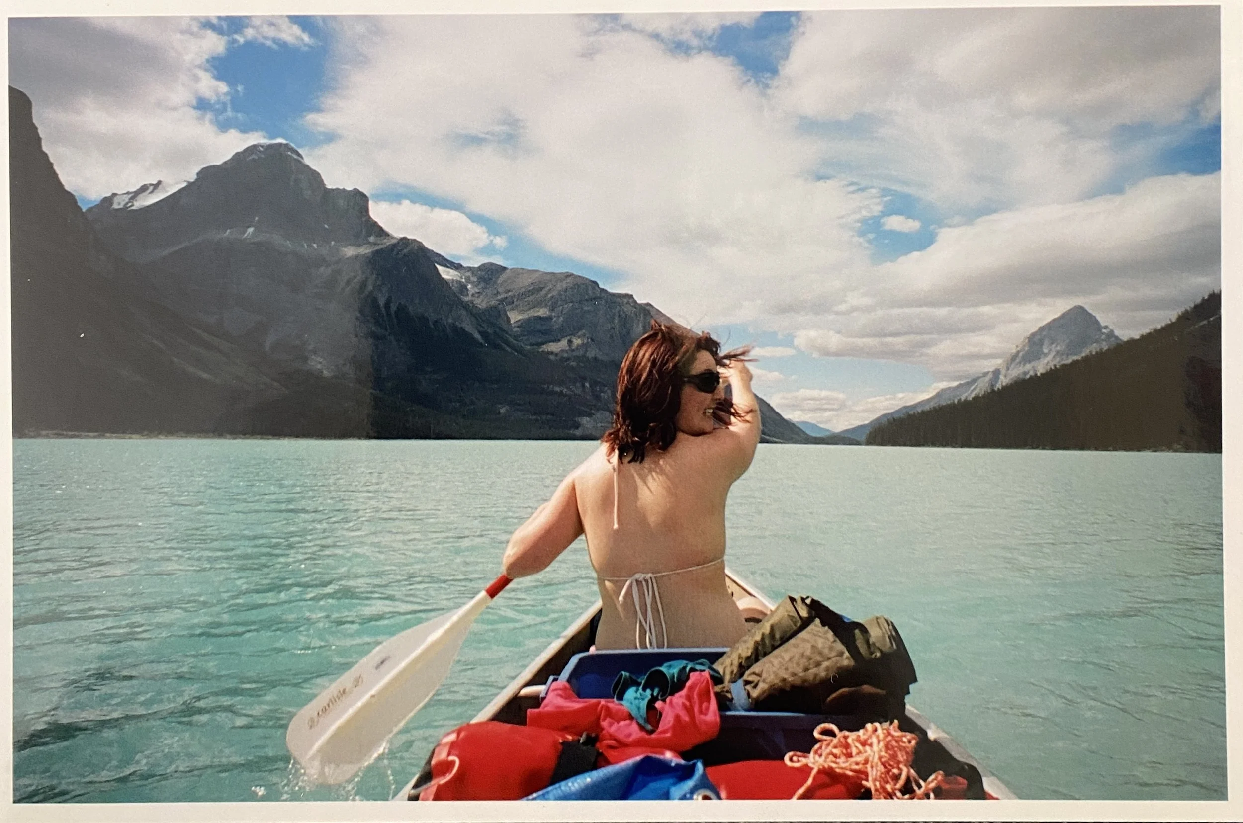 Sarah in the front of a canoe paddling on a huge green lake with mountains and blue sky around her