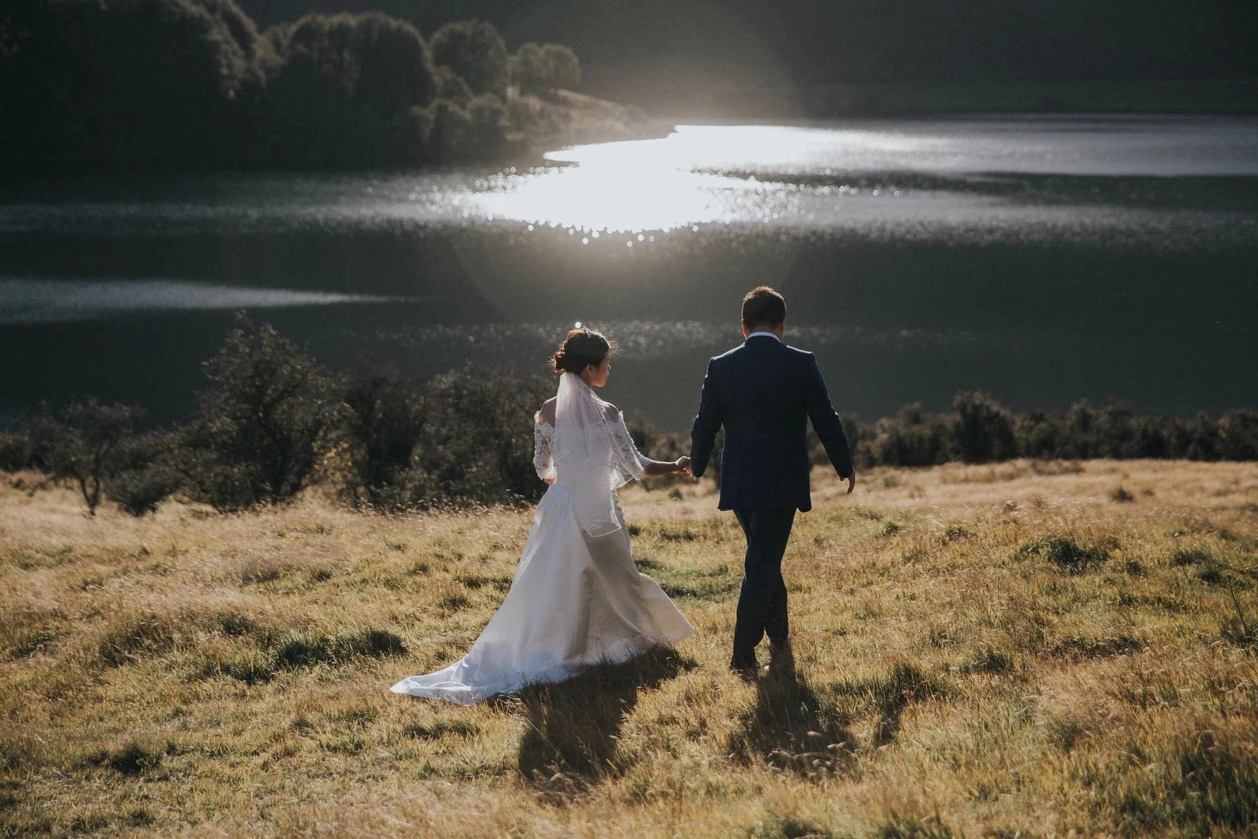 A bride and groom walking hand in hand in a grassy field with a lake and hills in the background during sunset.