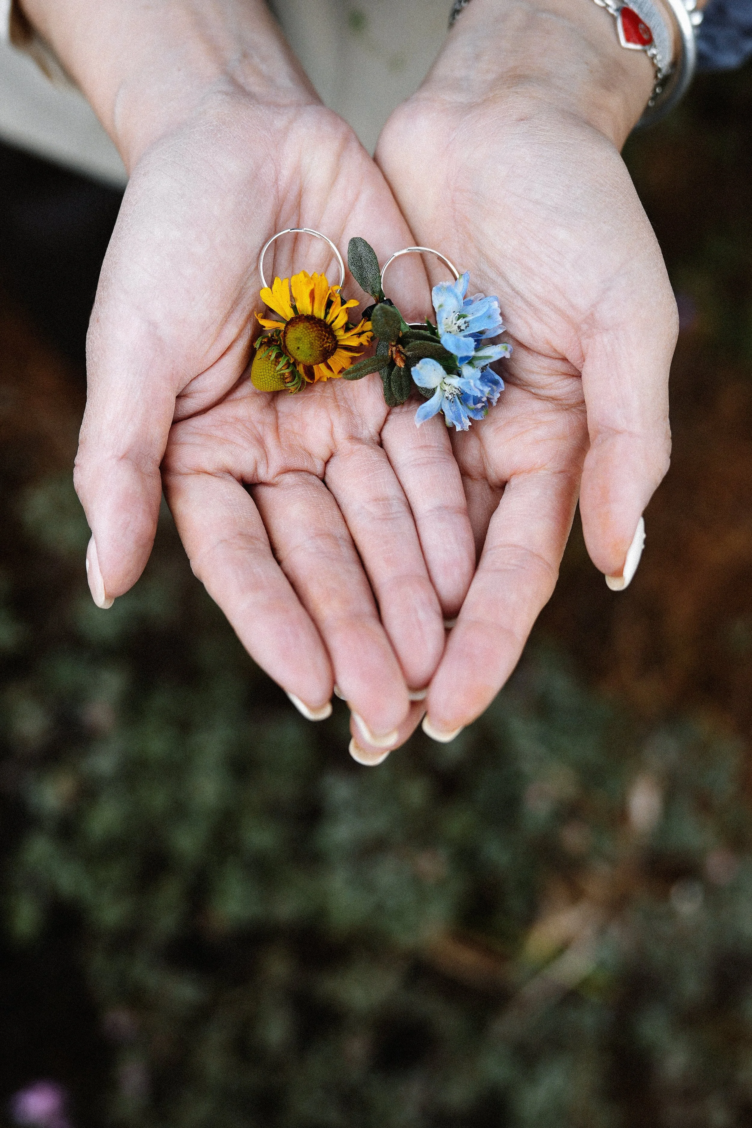 A pair of hands holding two wedding rings decorated with small flowers, one with a yellow flower and the other with blue flowers.