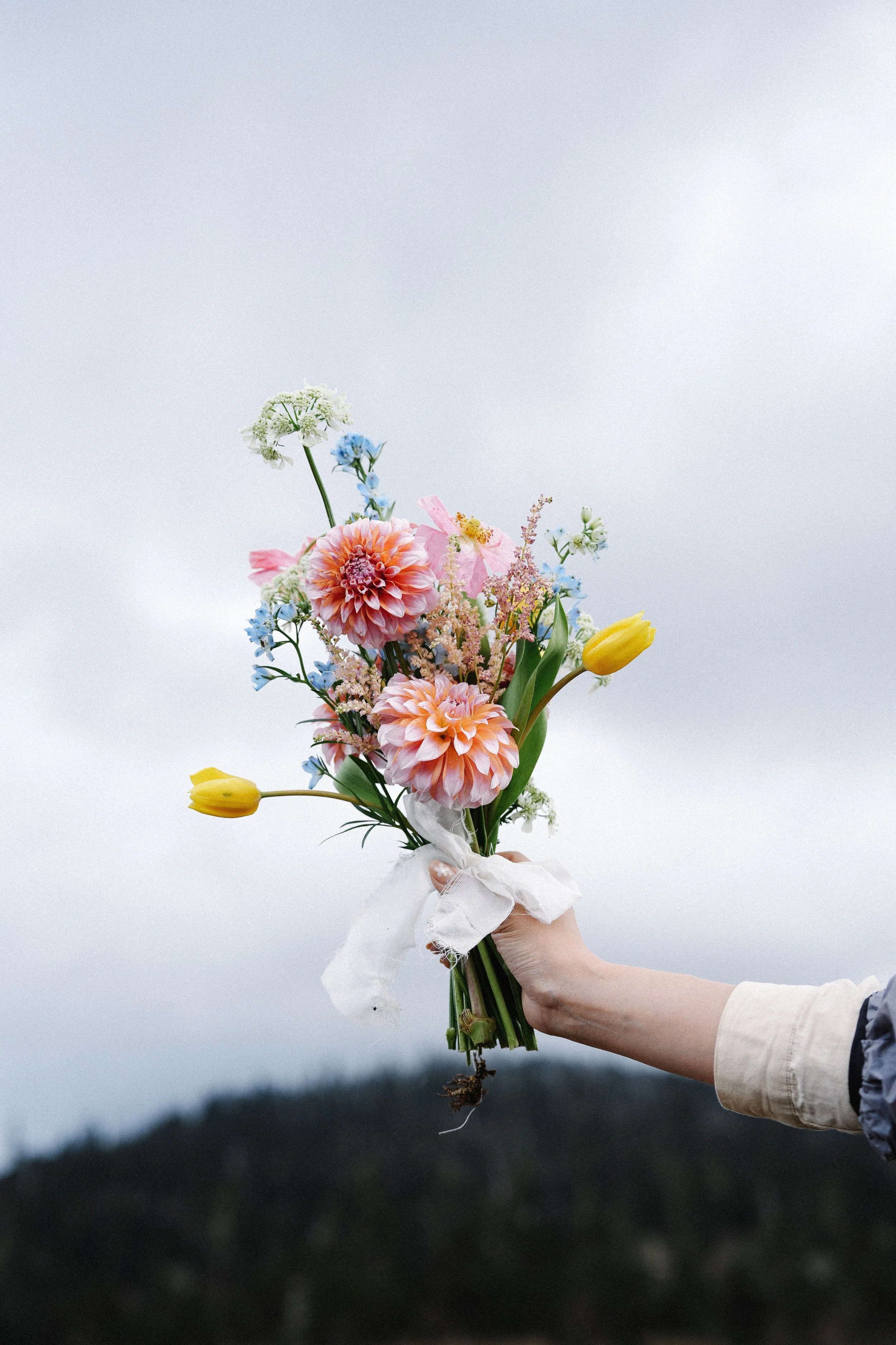 A hand holding a bouquet of colorful flowers against a cloudy sky.