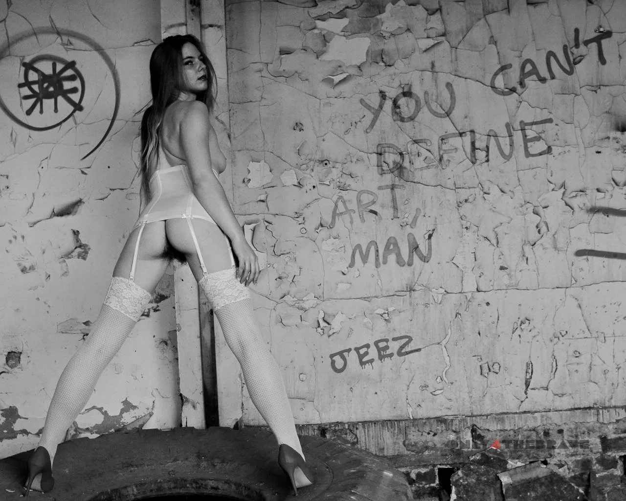 A woman in lingerie, stockings, and high heels posing against a distressed wall with graffiti that reads 'You can't define art, man' and a symbol in a circle.