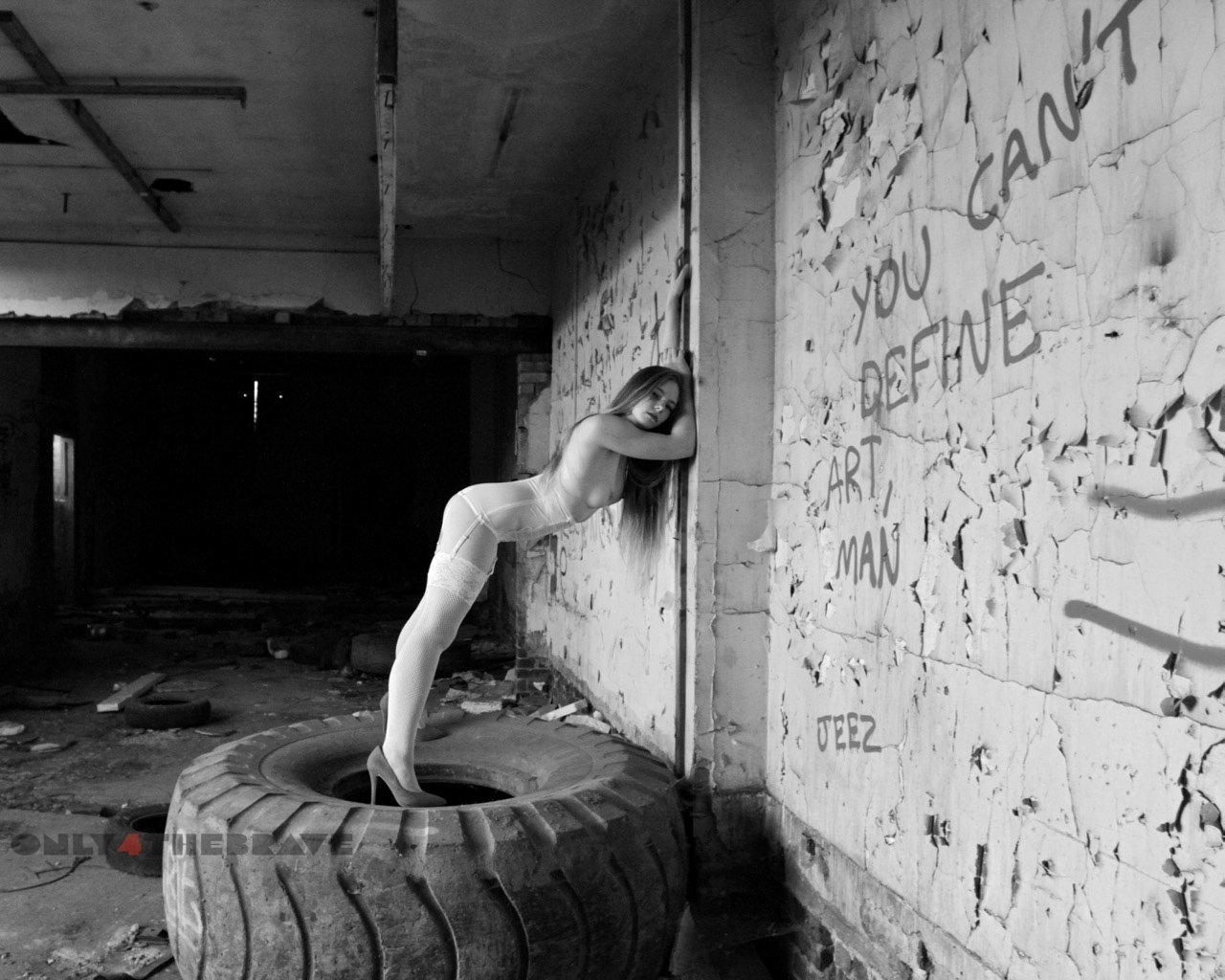 A black and white photography of a woman in a derelict building leaning towards the wall while standing on a large tire.