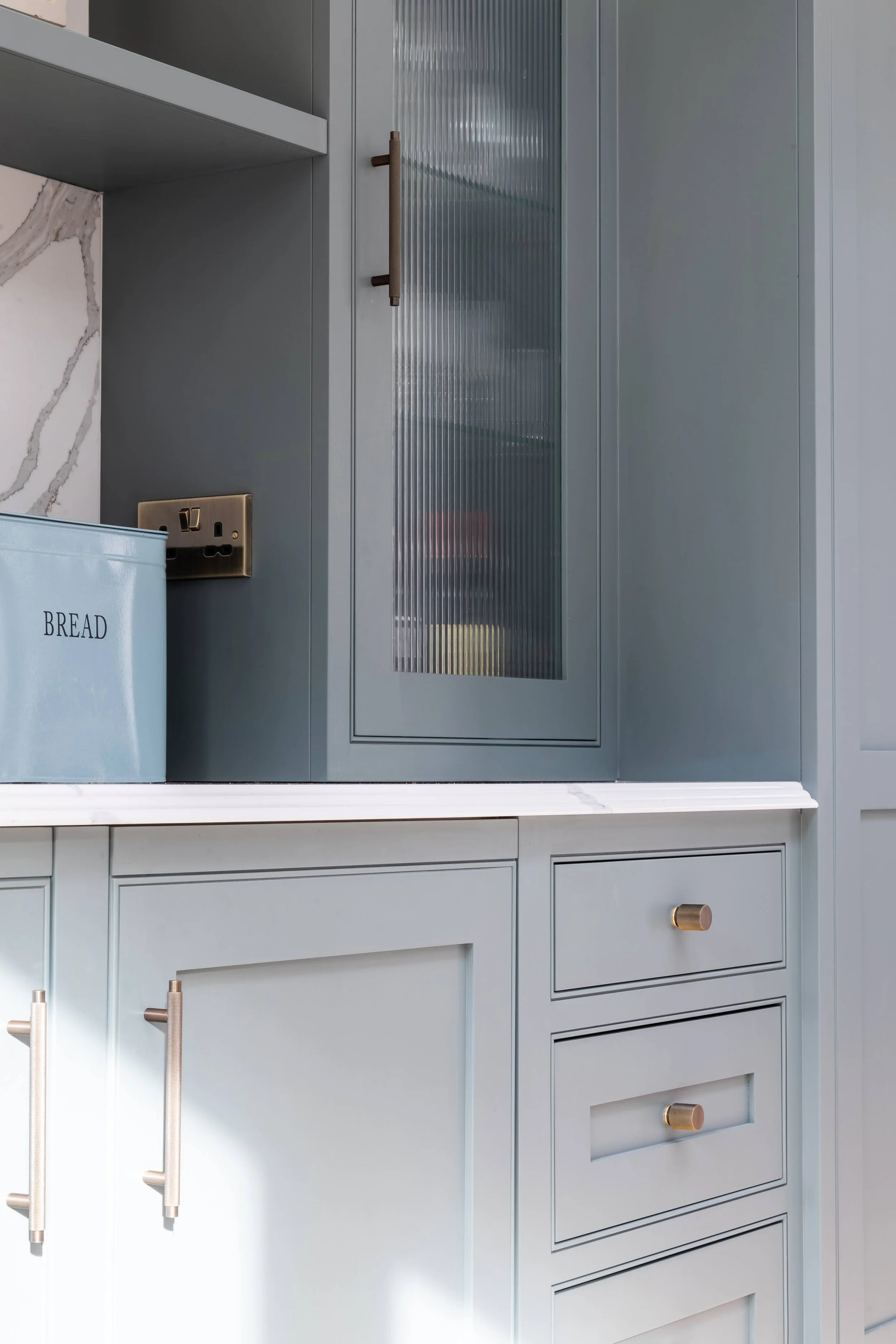Close-up view of a modern grey kitchen cabinet with gold-colored handles and a white countertop. A blue bread box is visible, along with a power outlet and a cabinet with a frosted glass door.