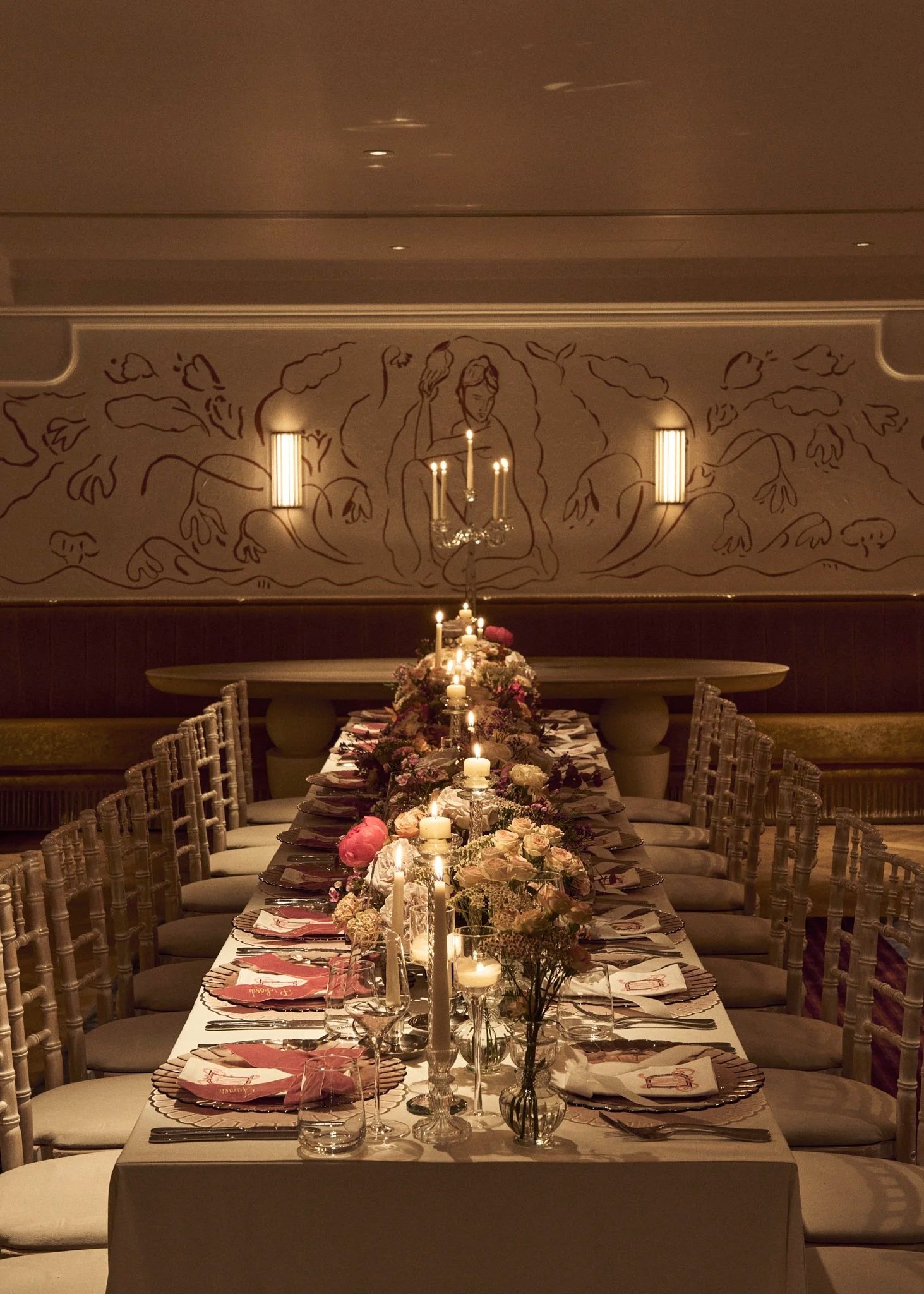A long banquet table decorated with candles, flowers, and place settings, set for a formal dinner in an elegant room with wall art and lighting.