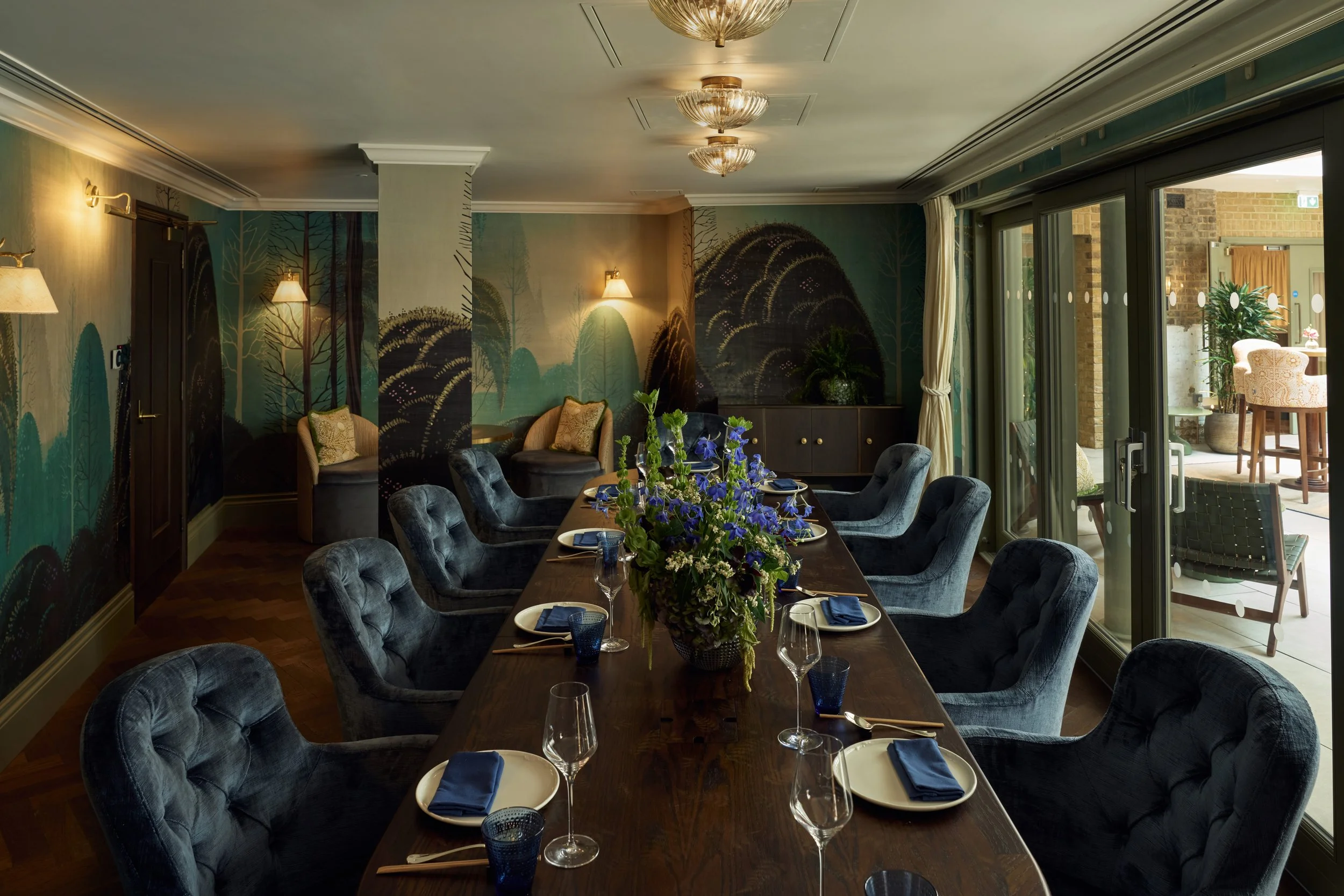 A dining room with a large wooden table set for a meal, surrounded by upholstered chairs. The table has a floral centerpiece, glassware, and napkins. The room features a mural with trees on the walls, wall-mounted lamps, and a sliding glass door leading to an outdoor patio with additional seating and plants.