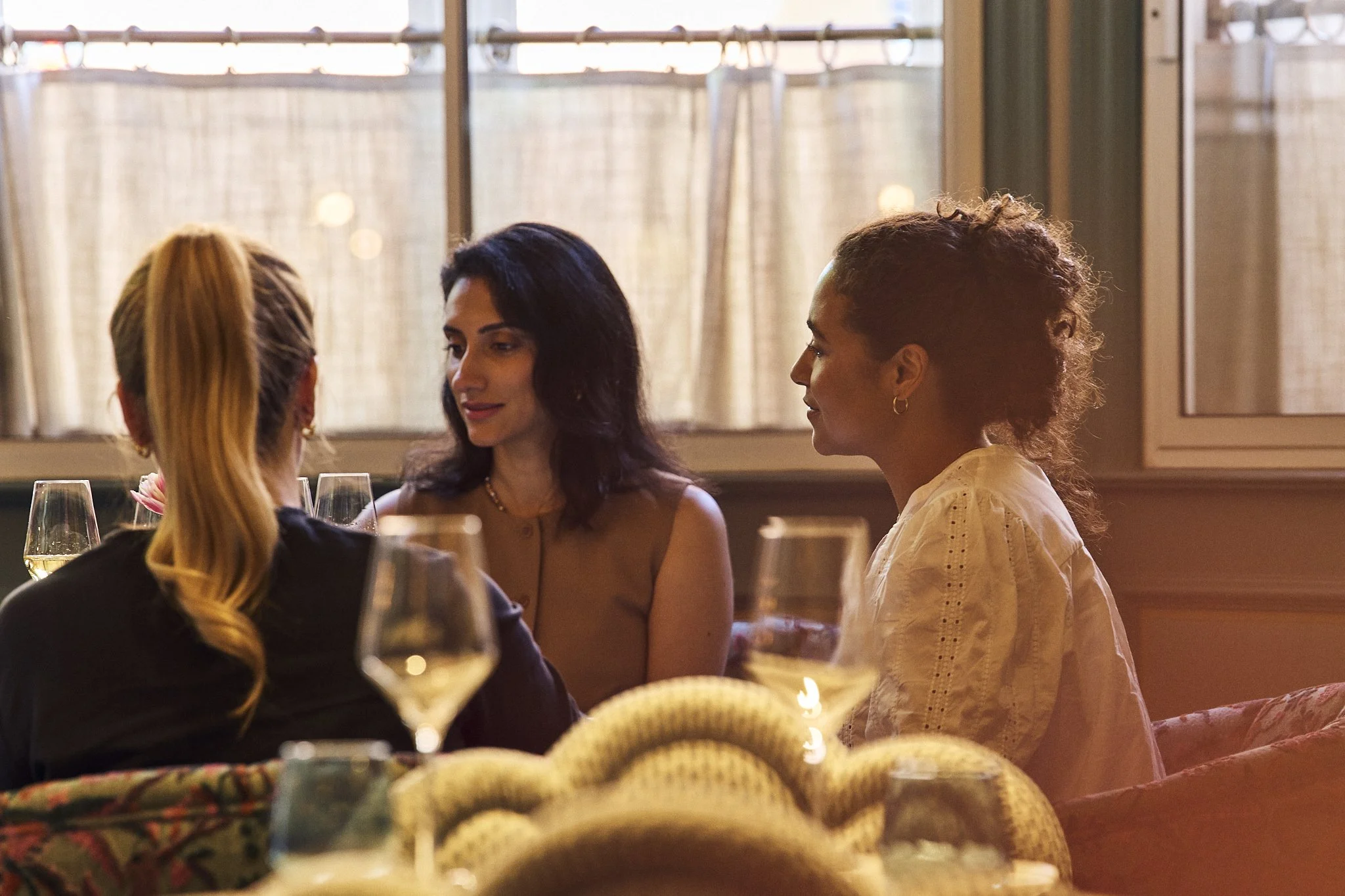 Group of women sitting at a dining table engaged in conversation, with wine glasses and a plate of bread in the foreground, in a warmly lit restaurant.