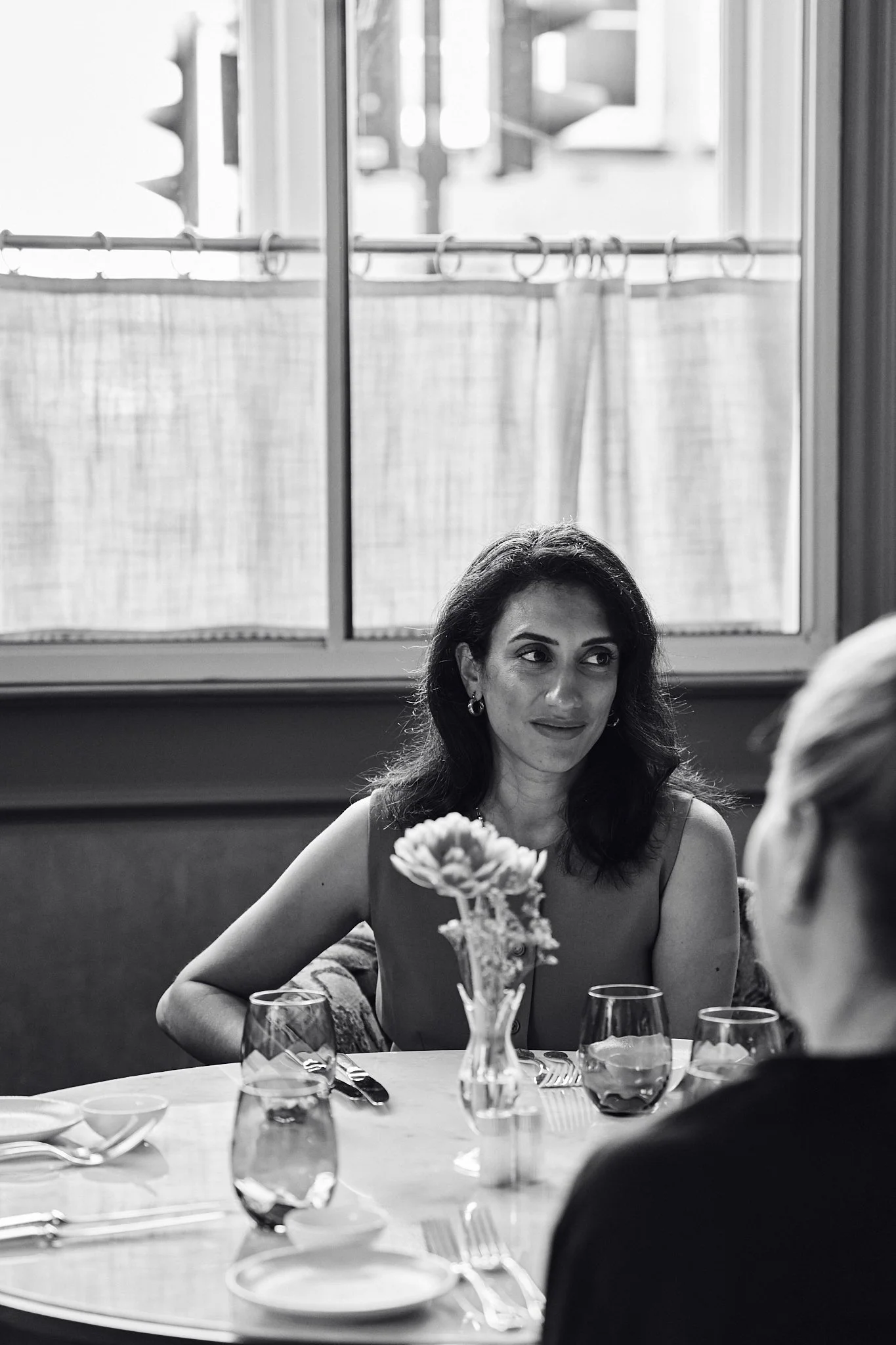 A woman with dark hair and earrings sitting at a table set for a meal. The table has a flower arrangement and wine glasses. The background shows a window with curtains and a laundry line outside.