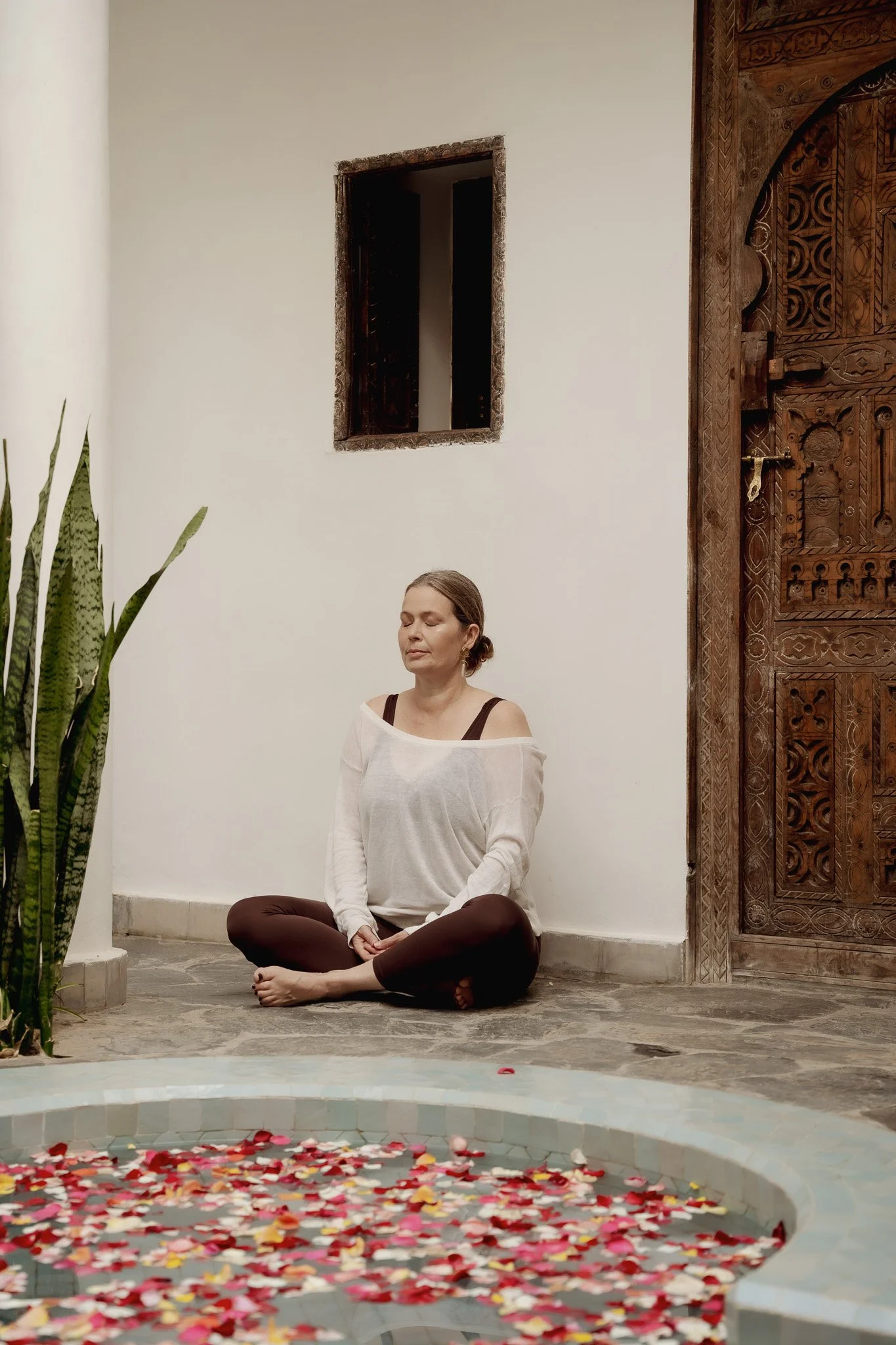 Een vrouw zit in een meditatieve pose met gesloten ogen, gekleed in een wit shirt en bruin leggings, zittend op de vloer naast een bad gevuld met water en rozenblaadjes, binnen een kamer met een houten deur en een cactusplant.