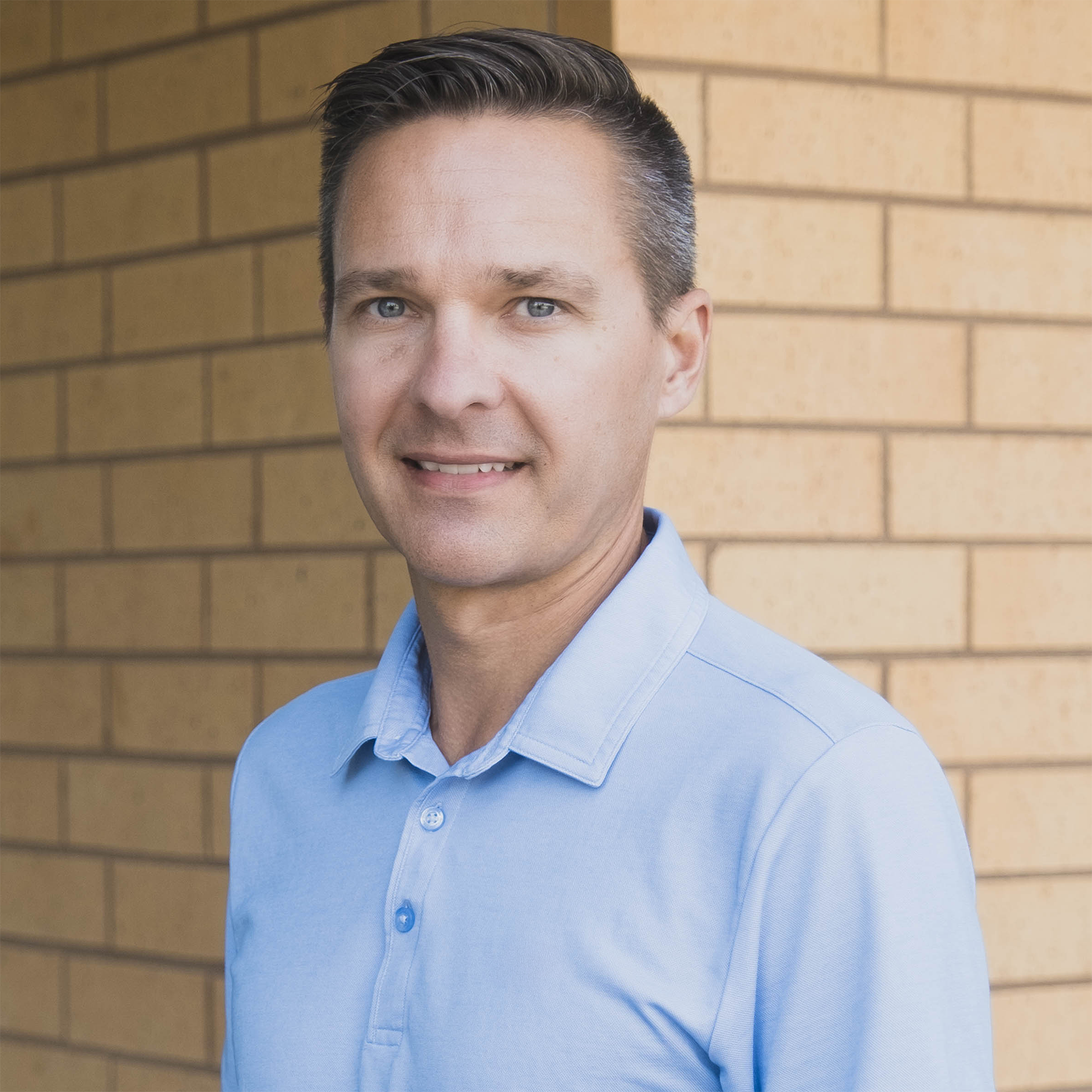 A man with short dark hair, wearing a light blue collared shirt, standing in front of a brick wall.