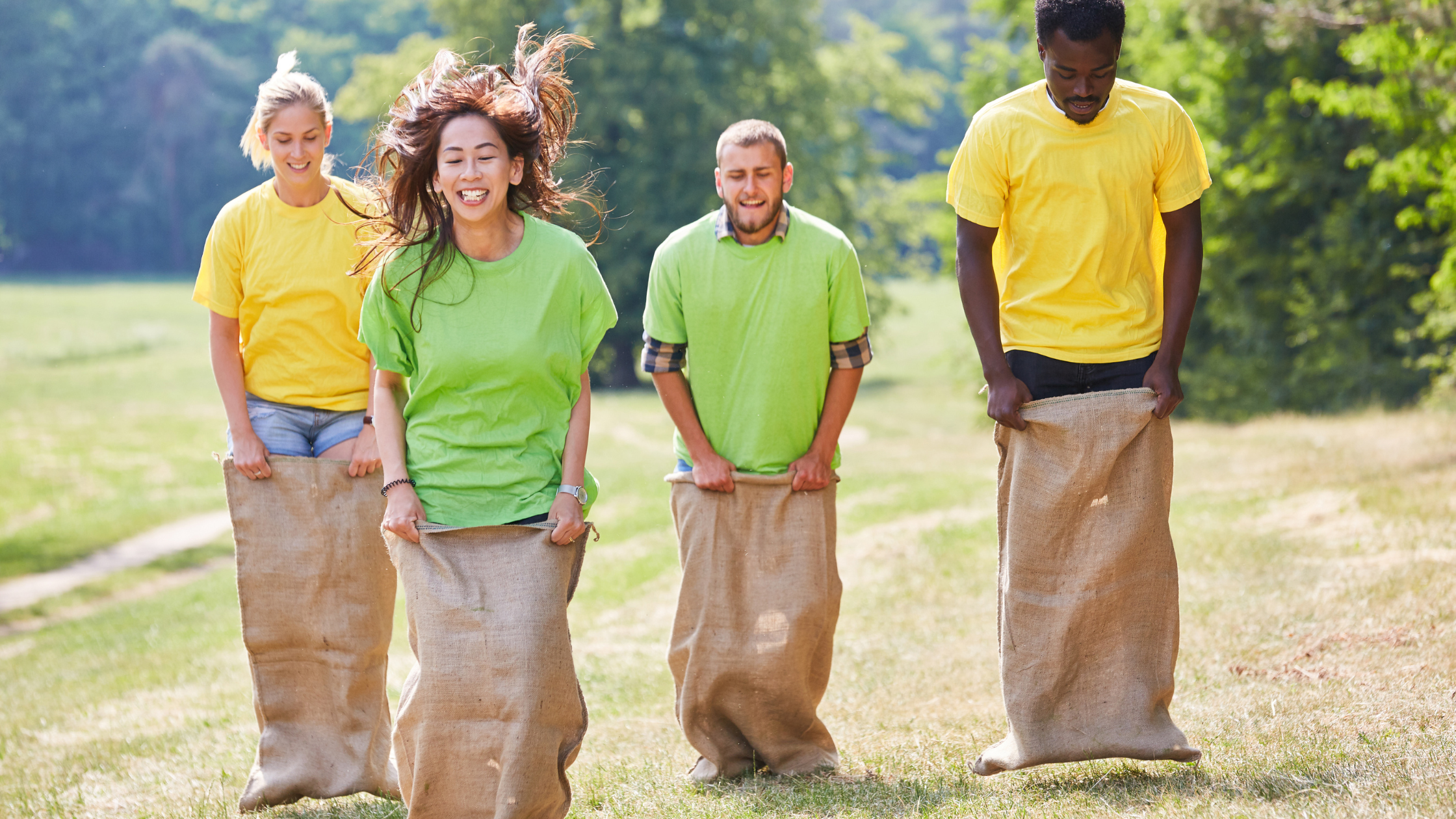 Employees wearing bright team shirts competing in a sack race during a company summer field day outdoors.