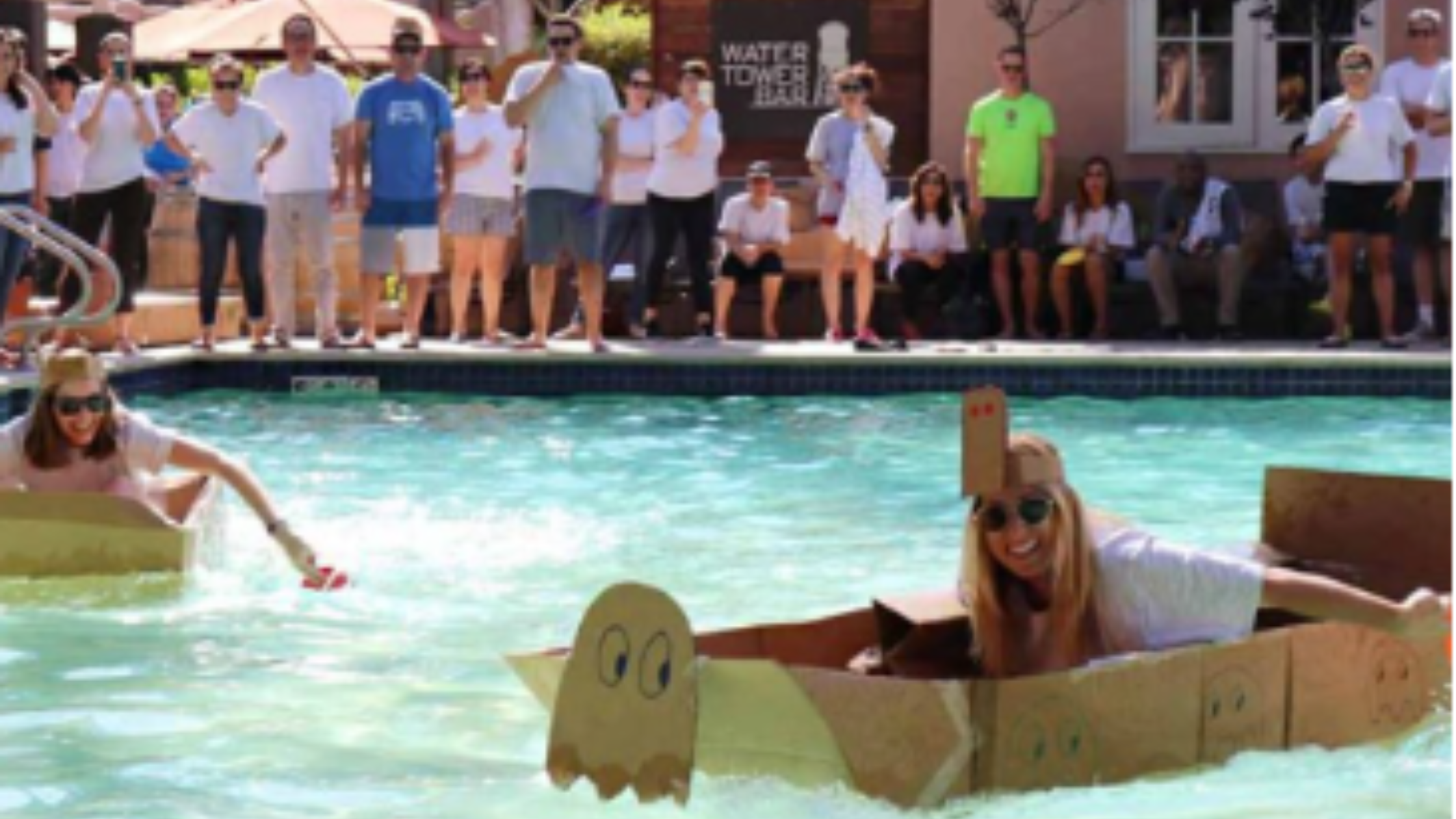 Employees racing handmade cardboard boats in a pool while coworkers cheer from the sidelines during a team summer challenge event.