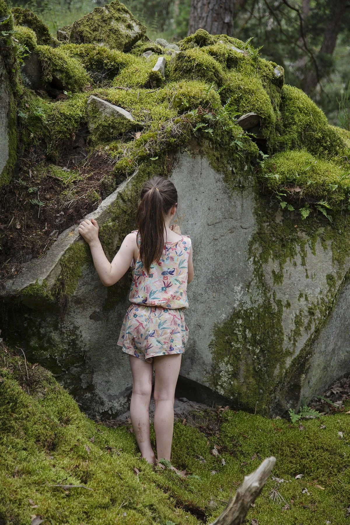 A young girl with brown hair in a ponytail, wearing a sleeveless floral top and shorts, standing barefoot on mossy ground, holding onto a moss-covered large rock in a forest.