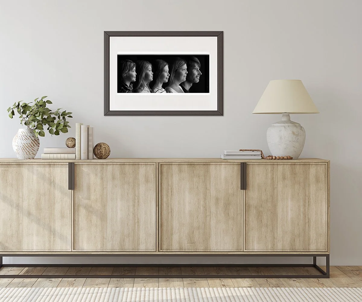 Black and white family portrait framed on a wall above a buffet with a lamp, vase and books.