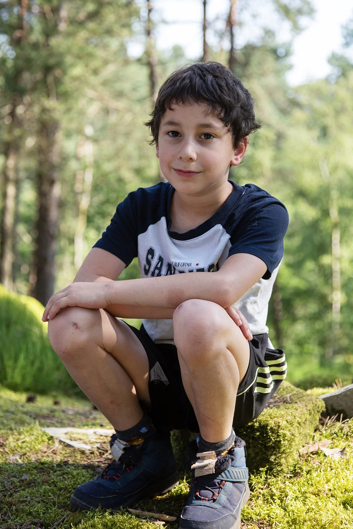 A young boy with short dark hair and a bruise on his knee sitting on a moss-covered log in a forest, wearing a black and white T-shirt and black shorts with yellow stripes.