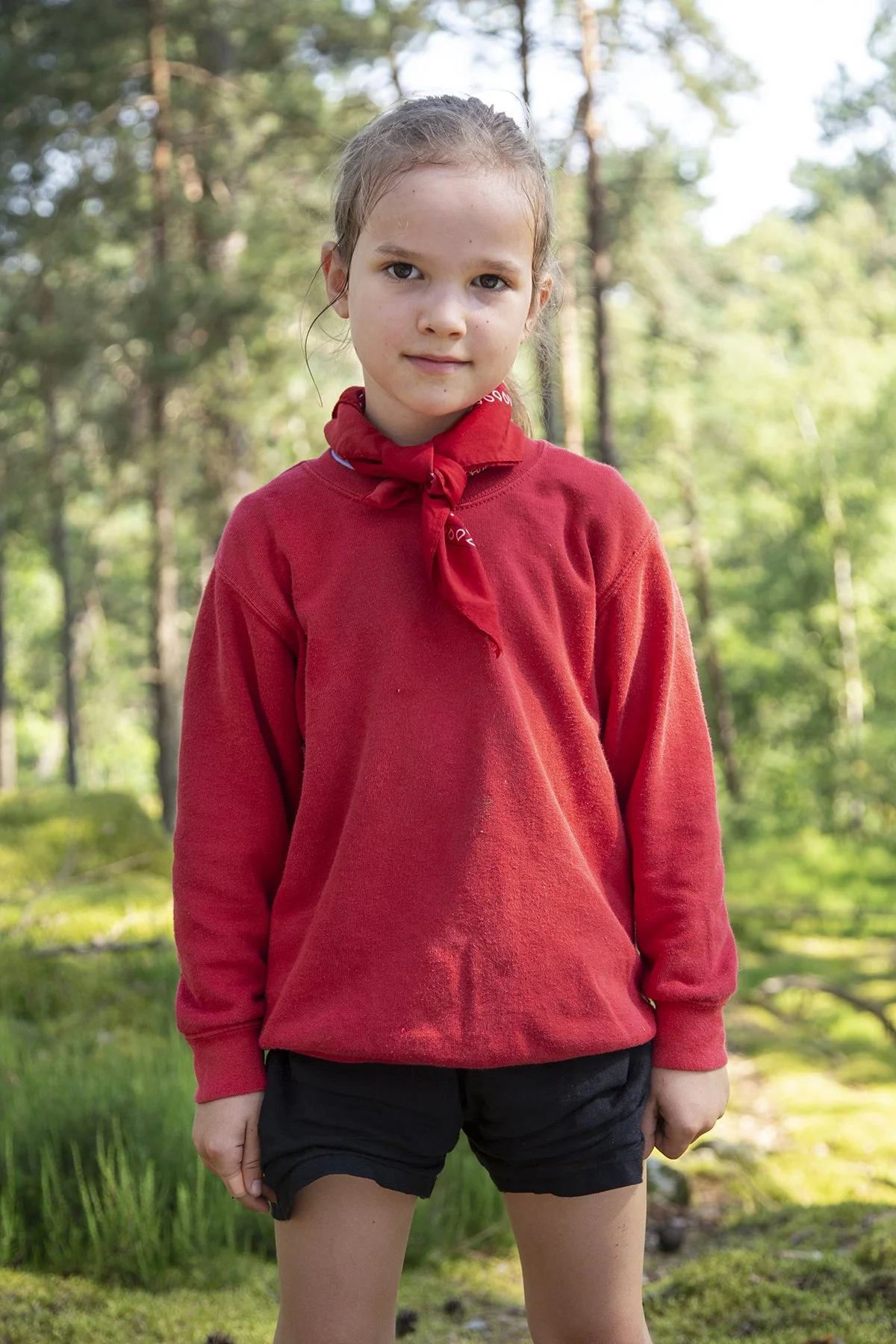 A young girl in a red long-sleeve shirt with a red neckerchief stands outdoors in a forested area.