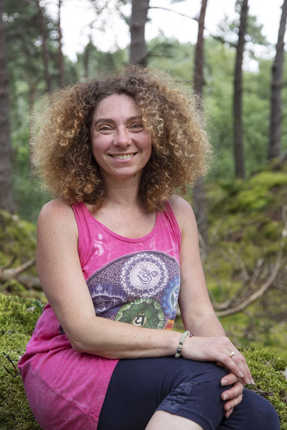 A woman with curly blonde hair smiling and sitting in a lush green forest, wearing a pink sleeveless top with a colorful mandala design and black pants.