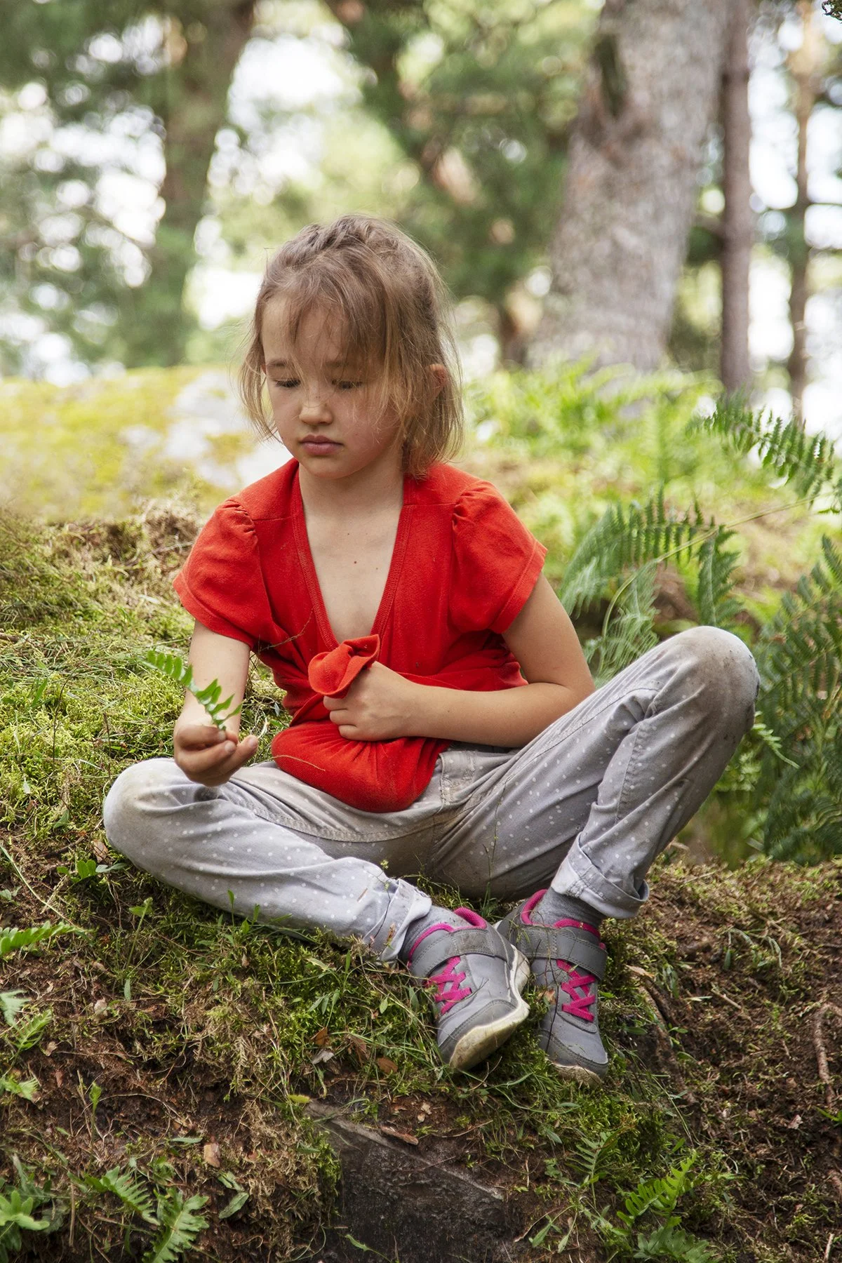 A young girl with light skin, brown hair, and a somber expression sitting on the grassy ground in a forest, holding a small green fern leaf in her hand, surrounded by trees and lush green foliage.