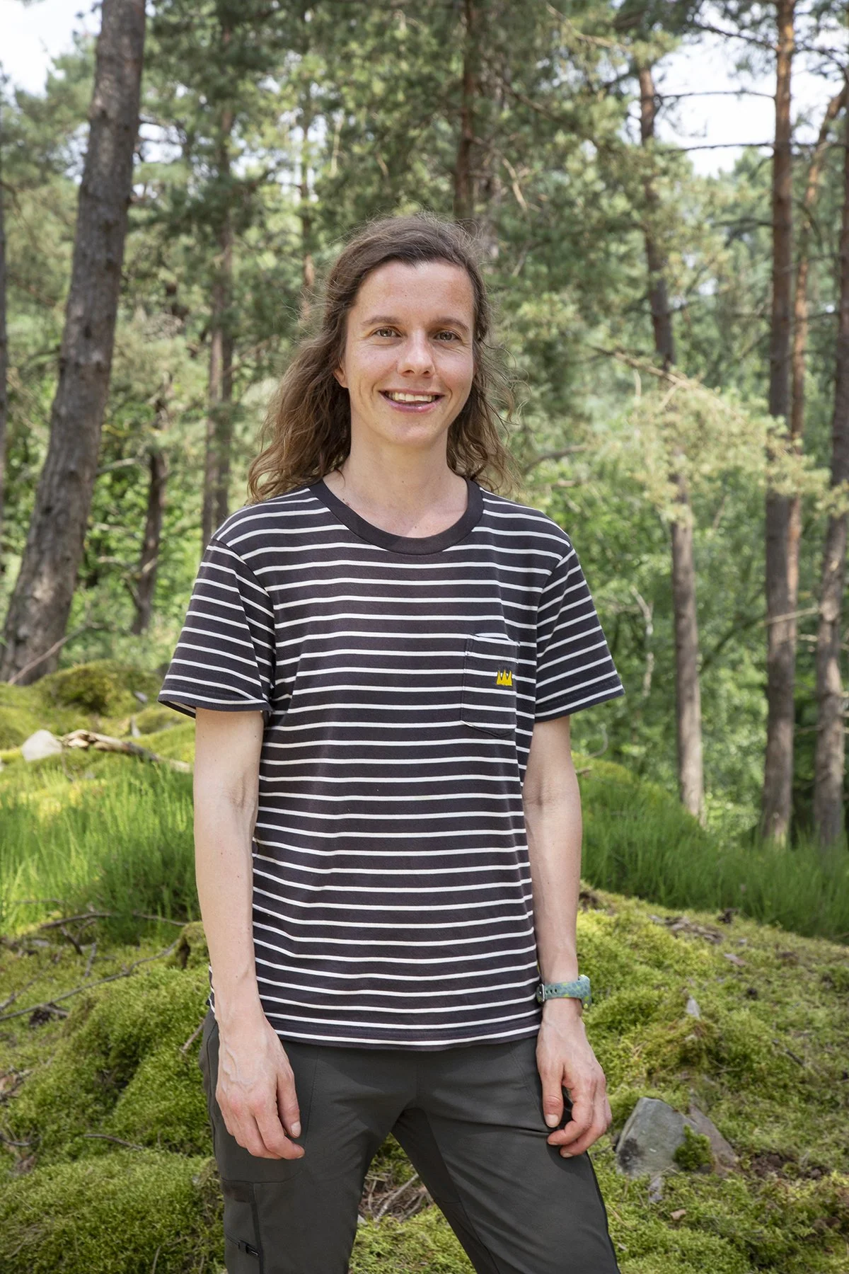 A woman standing outdoors in a forested area with green trees and mossy ground, wearing a black and white striped t-shirt with a small yellow crown emblem on the chest and dark pants.