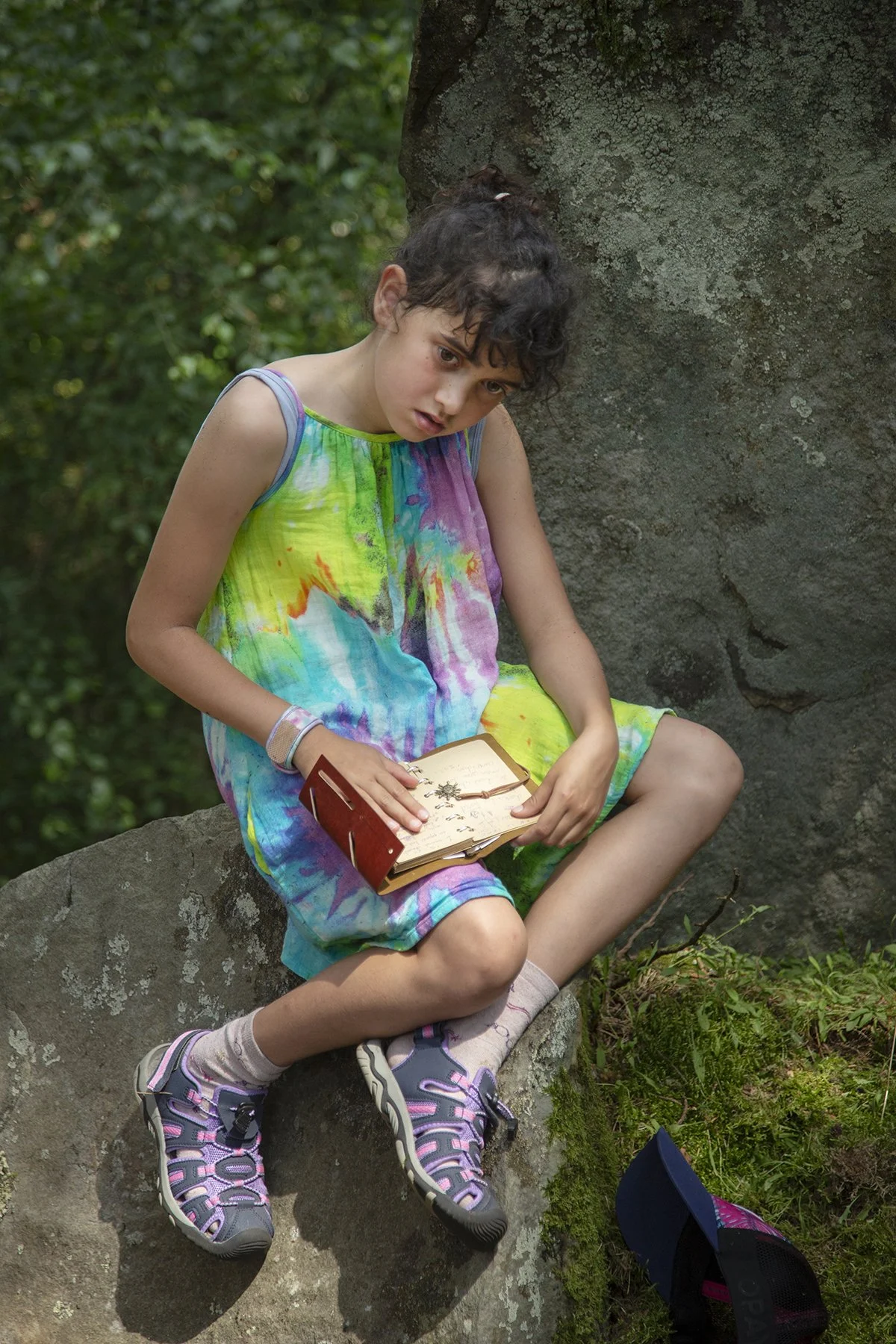 Young girl with curly hair sitting on a rock outdoors, holding an open book, wearing a colorful tie-dye dress and sneakers.