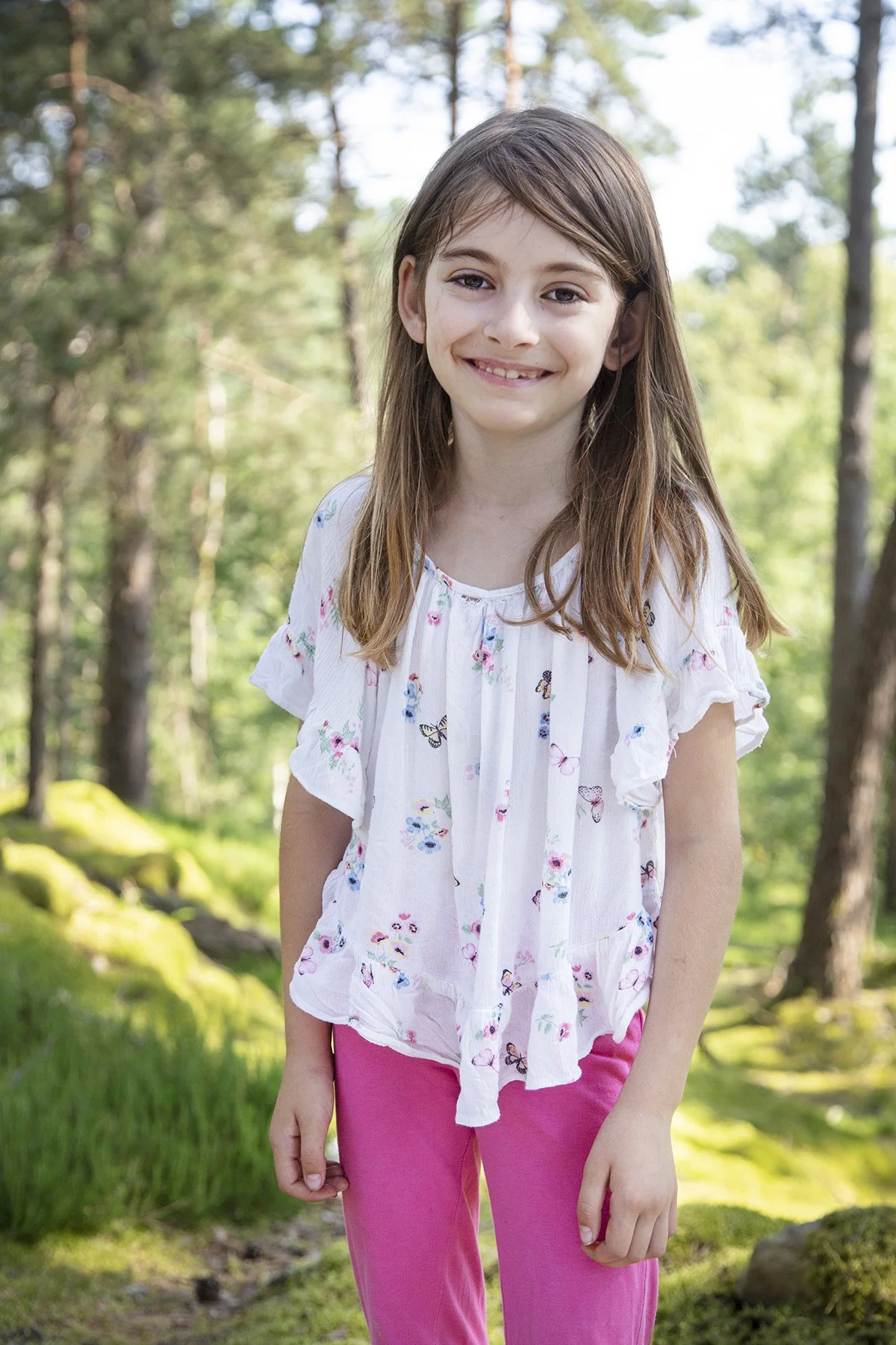 A young girl with long brown hair smiles at the camera while standing outdoors in a wooded area with trees and green mossy ground.