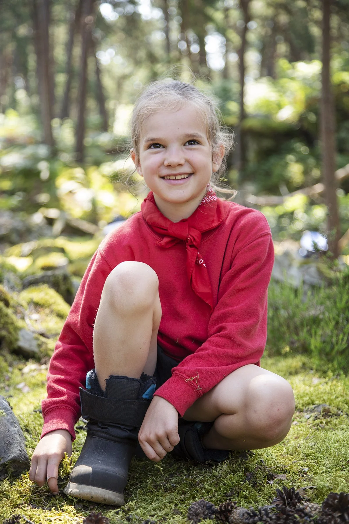 A young girl with blonde hair and a red bandana around her neck sitting on mossy ground in a forest, smiling at the camera.