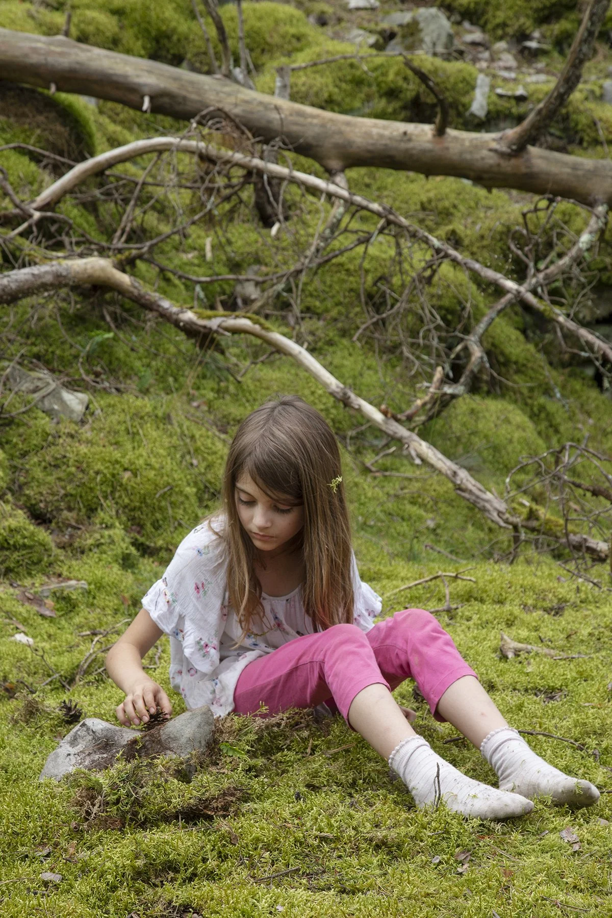 A young girl with brown hair and light skin sitting on mossy ground in a green forest with fallen branches and moss-covered rocks.