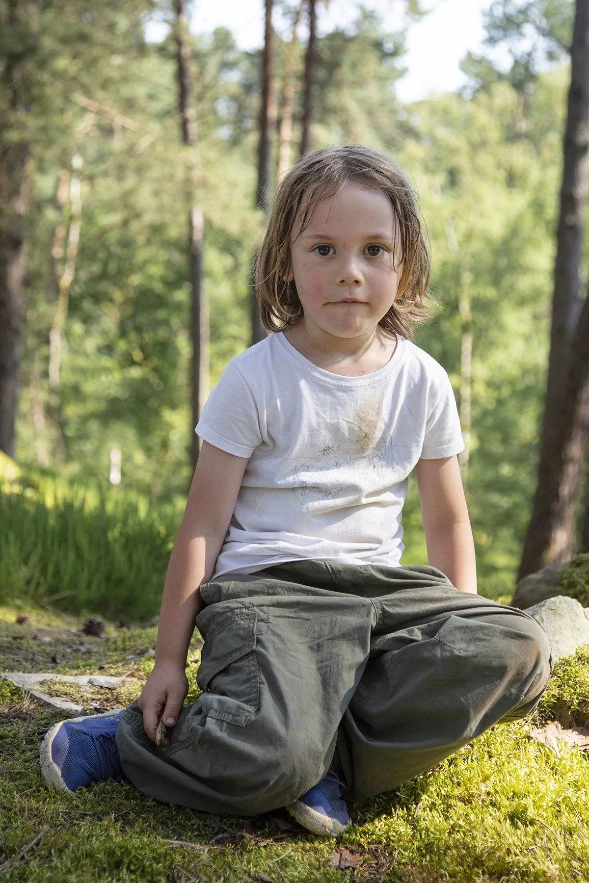 A young boy with long, wet hair sitting outdoors on a mossy ground in a forested area, surrounded by tall trees and green foliage.