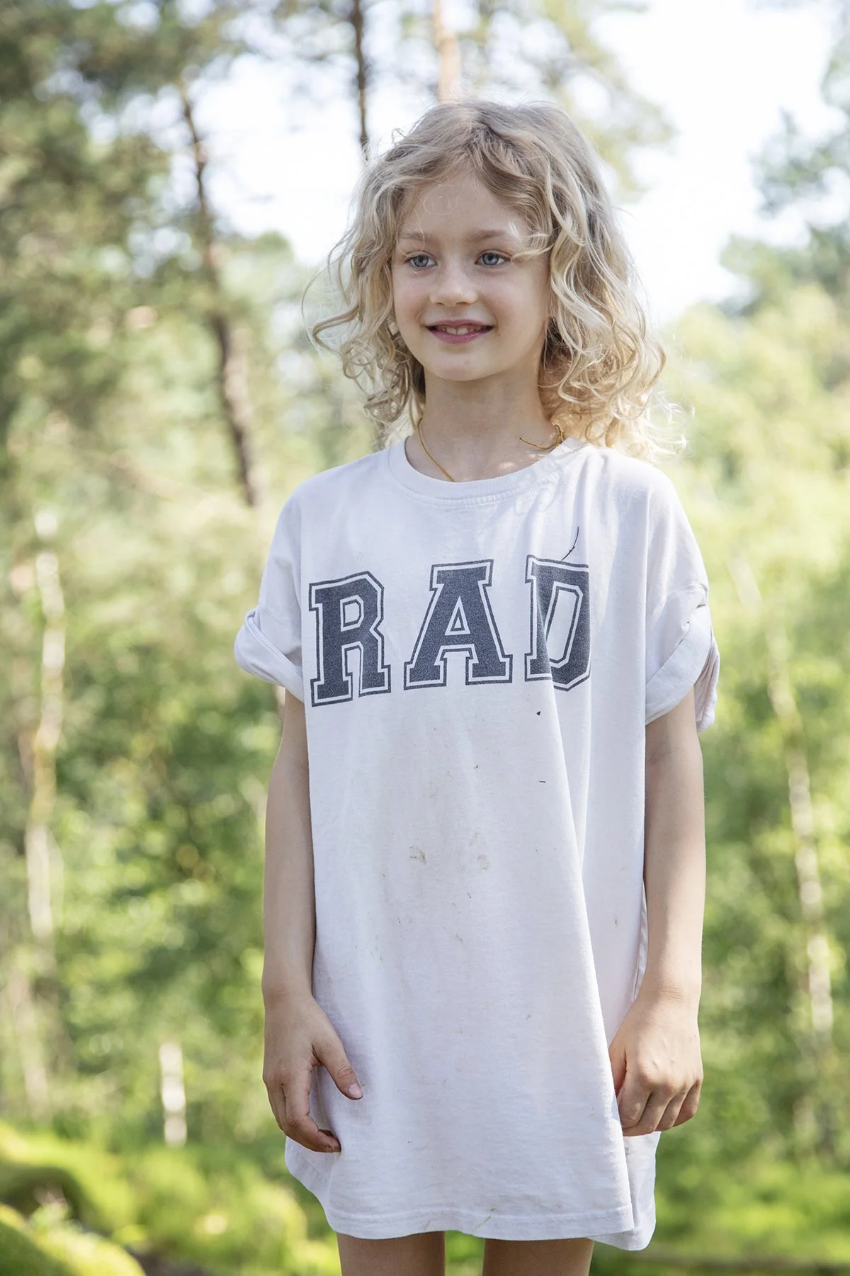 A young girl with curly blonde hair smiling outdoors in a forested area, wearing a white T-shirt with the word 'RAD' written on it.