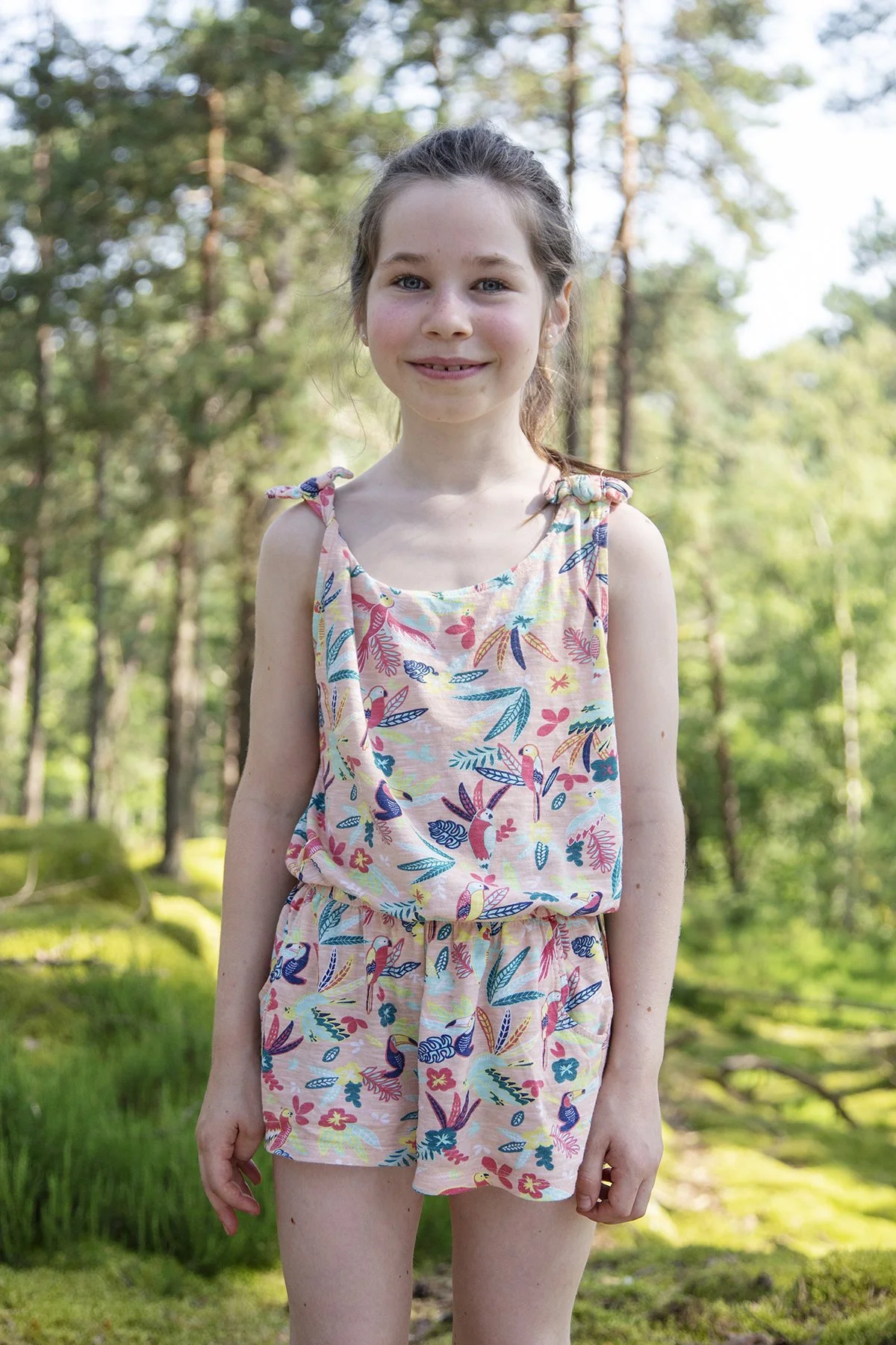 A young girl with long brown hair in a ponytail, smiling, standing outdoors in a wooded area with greenery and trees in the background, wearing a pink sleeveless top and shorts with a colorful bird and floral pattern.