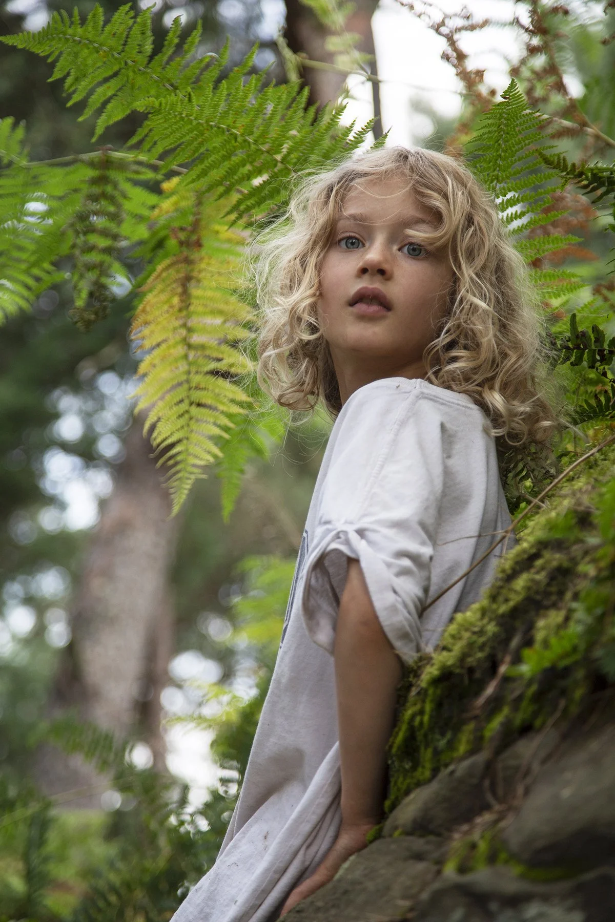 A young girl with curly blonde hair and blue eyes standing outdoors in a lush, green forested area. She is leaning against a moss-covered tree trunk, wearing a loose white shirt with rolled-up sleeves, and looking slightly over her shoulder with a cu