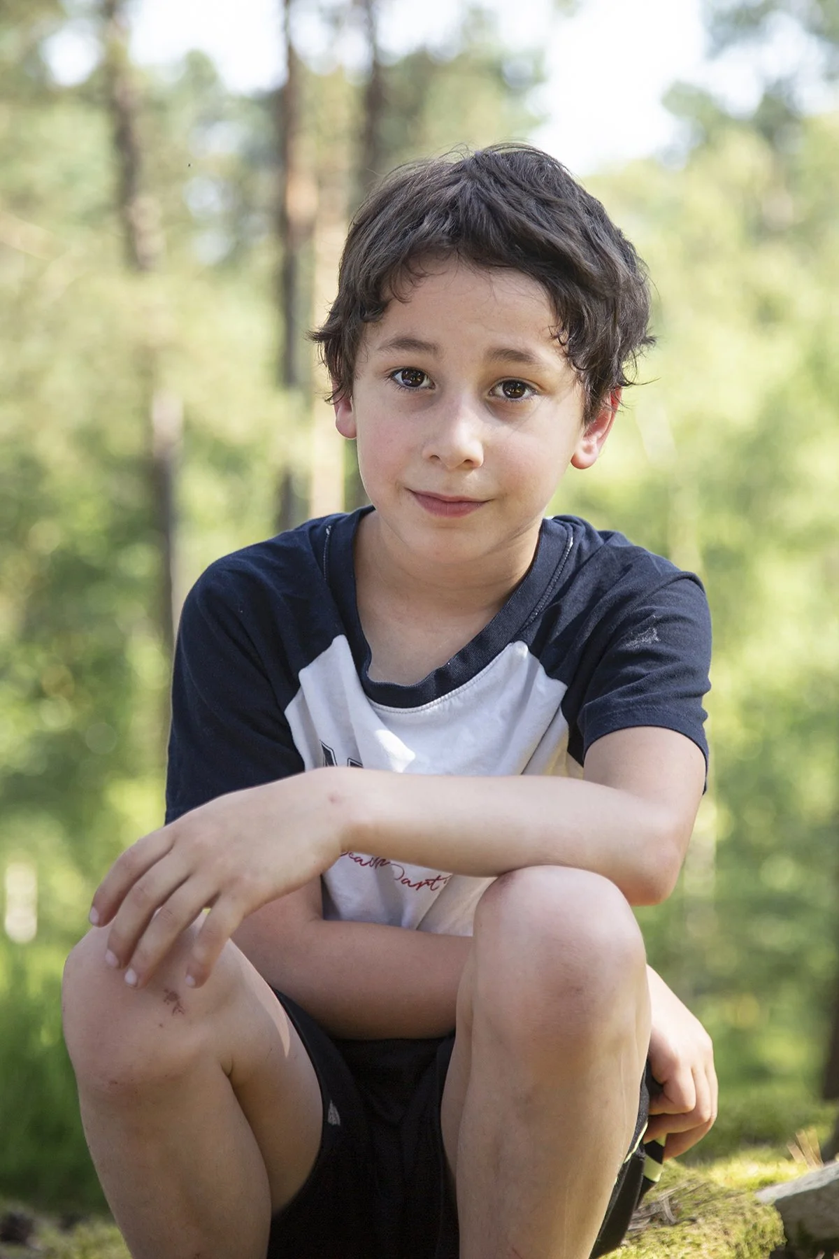 A young boy with brown hair and light skin sitting outdoors in a wooded area, wearing a black and white t-shirt and black shorts, with dirt and scrapes on his knees and arm, looking at the camera with a slight smile.