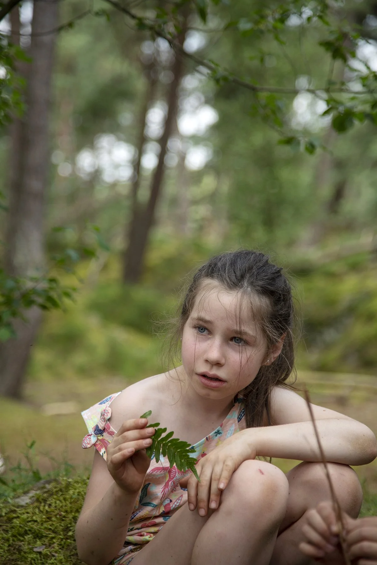 A young girl with long brown hair and blue eyes sitting on mossy ground in a forest, holding a green fern leaf, wearing a colorful patterned sleeveless dress, looking thoughtful against a background of trees and foliage.