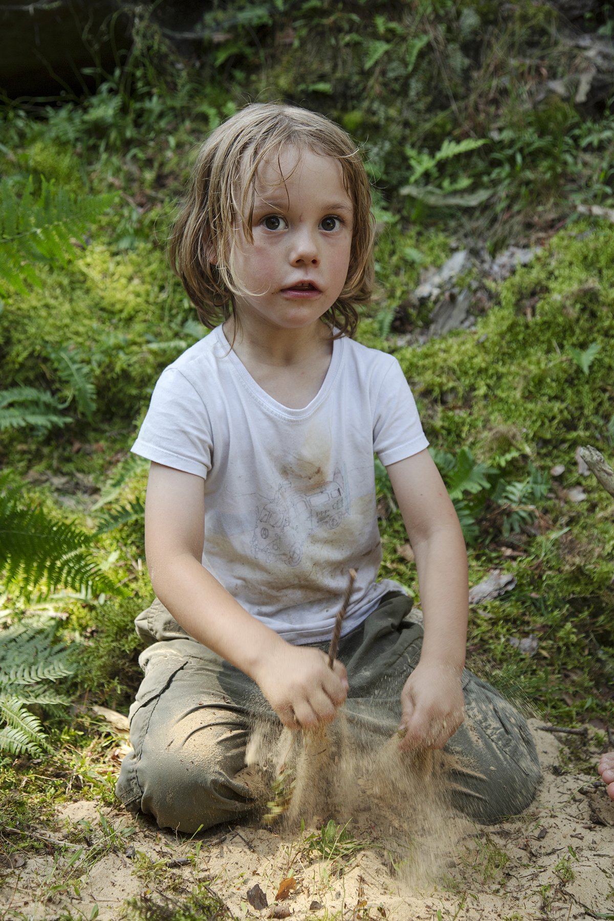 A young child with long, light brown hair and a white t-shirt is sitting on the sandy ground outdoors, playing with dirt and grass in a lush, green forest setting.
