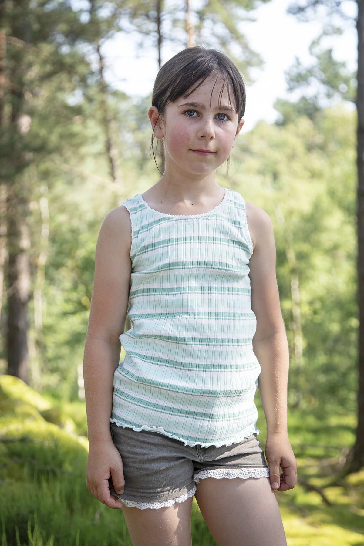 A young girl with brown hair and blue eyes standing outdoors in a wooded area, wearing a sleeveless striped top and gray shorts with lace trim.