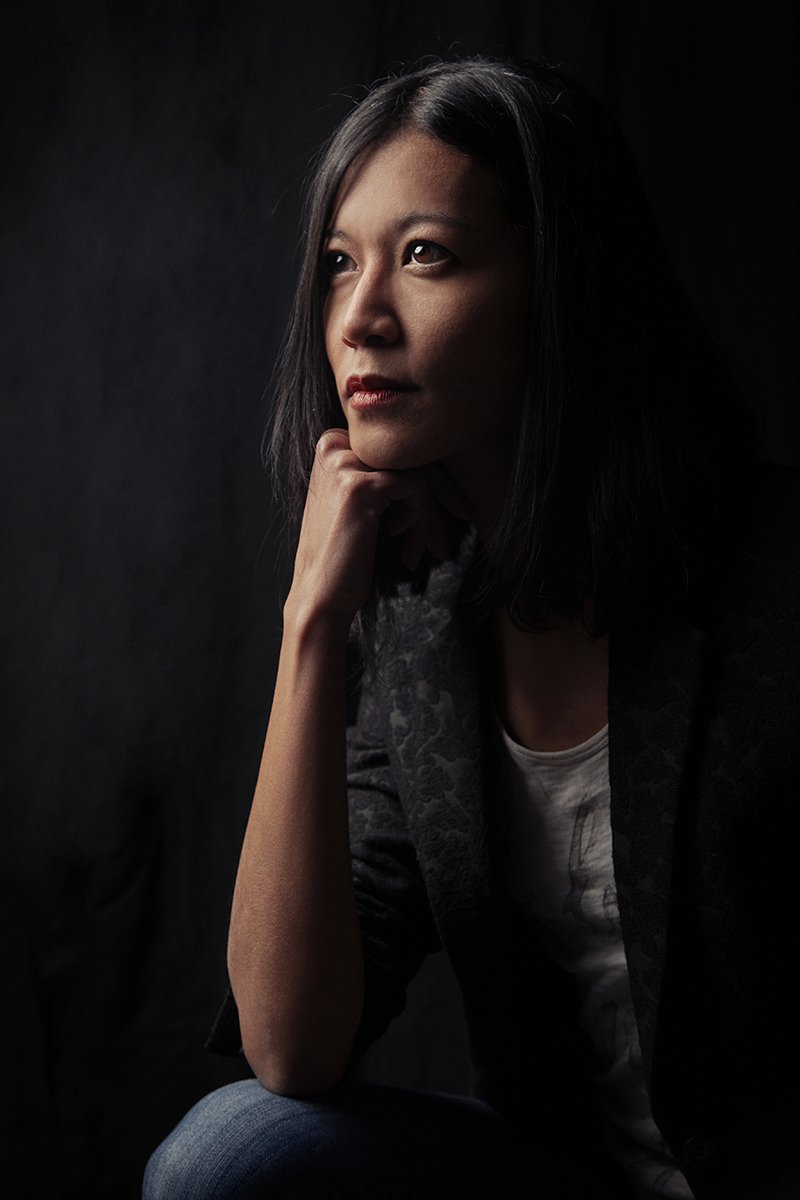 Young Vietnamese woman in semi-profile, soft light on her face, wearing black jacket against black background