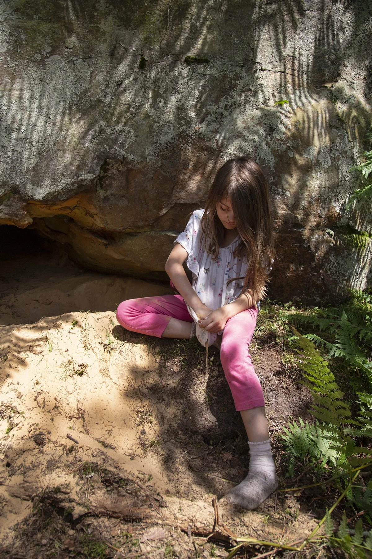 A young girl sitting on the dirt ground near a large rock with moss and trees casting shadows, wearing a light-colored shirt and pink pants, with her hair down, looking down and holding her lap.