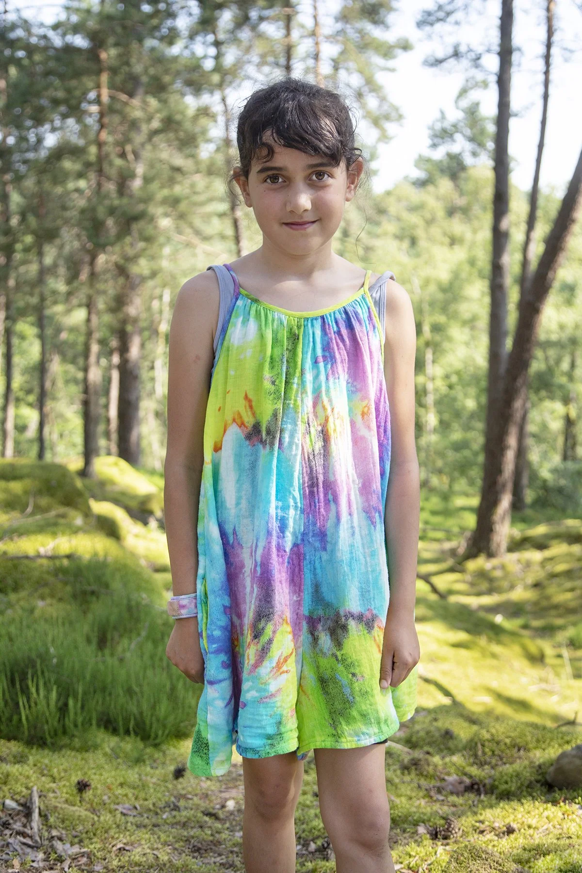 A young girl standing in a lush green forest with tall trees in the background, wearing a colorful tie-dye dress.
